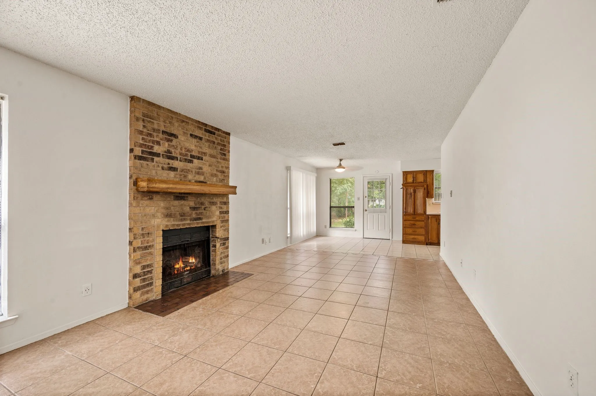 Brick fireplace with wood mantel, tile flooring, white walls, and a ceiling fan