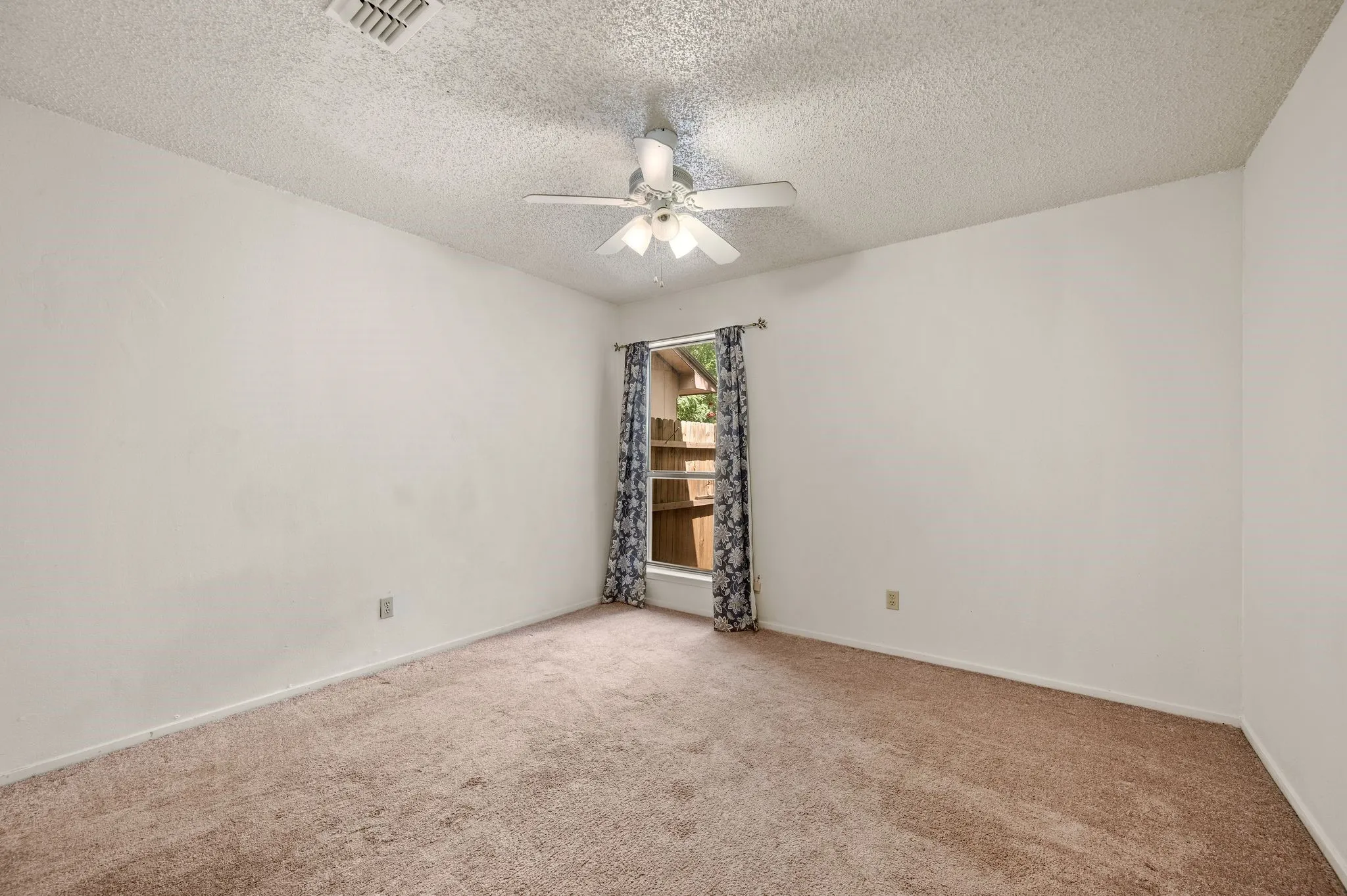 Carpeted flooring, white painted walls, window with curtains, and a ceiling fan with integrated lighting