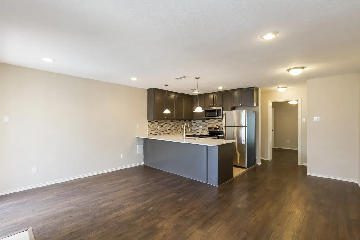 Open-concept living and kitchen area featuring dark cabinetry with pendant lighting, stainless steel appliances, geometric tile backsplash, wood-finish flooring throughout, and recessed ceiling lighting