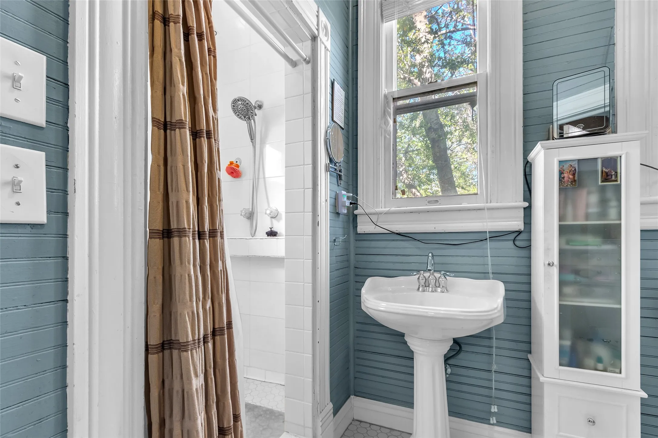 Bathroom with white tile shower, porcelain sink, wooden cabinetry, natural light from window, and a beige shower curtain visible