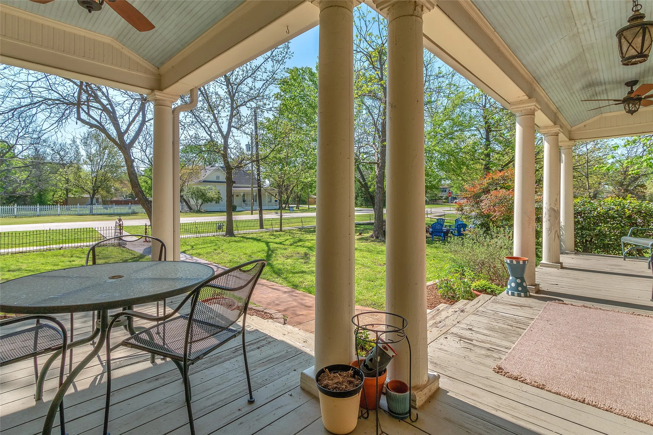 Covered front porch with circular metal table, four chairs, wooden flooring, and sunlight filtering through trees in a landscaped yard.