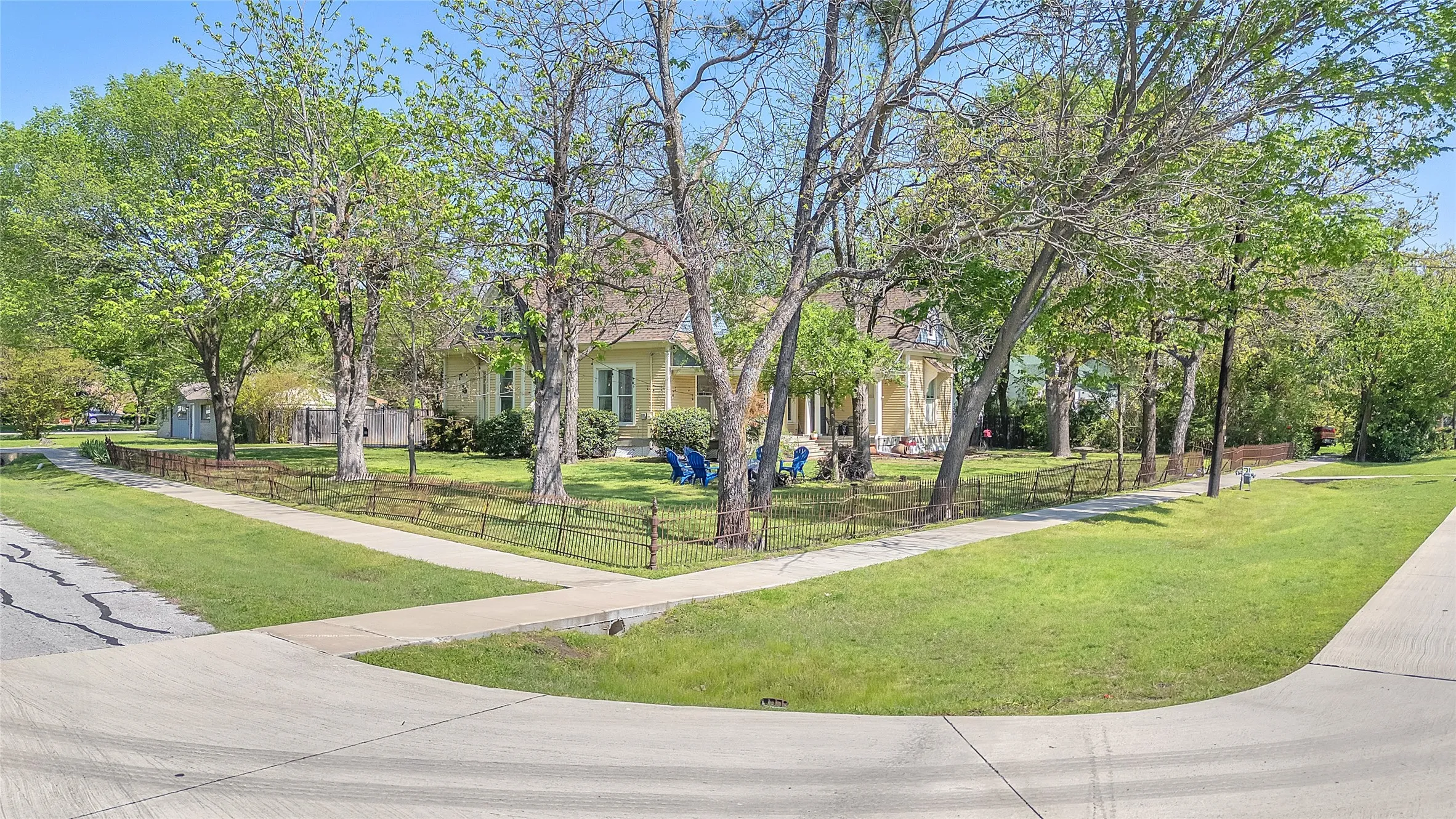 Front yard with landscaped grass, mature trees, and iron fencing surrounding a single-story house with light-colored siding and a covered porch
