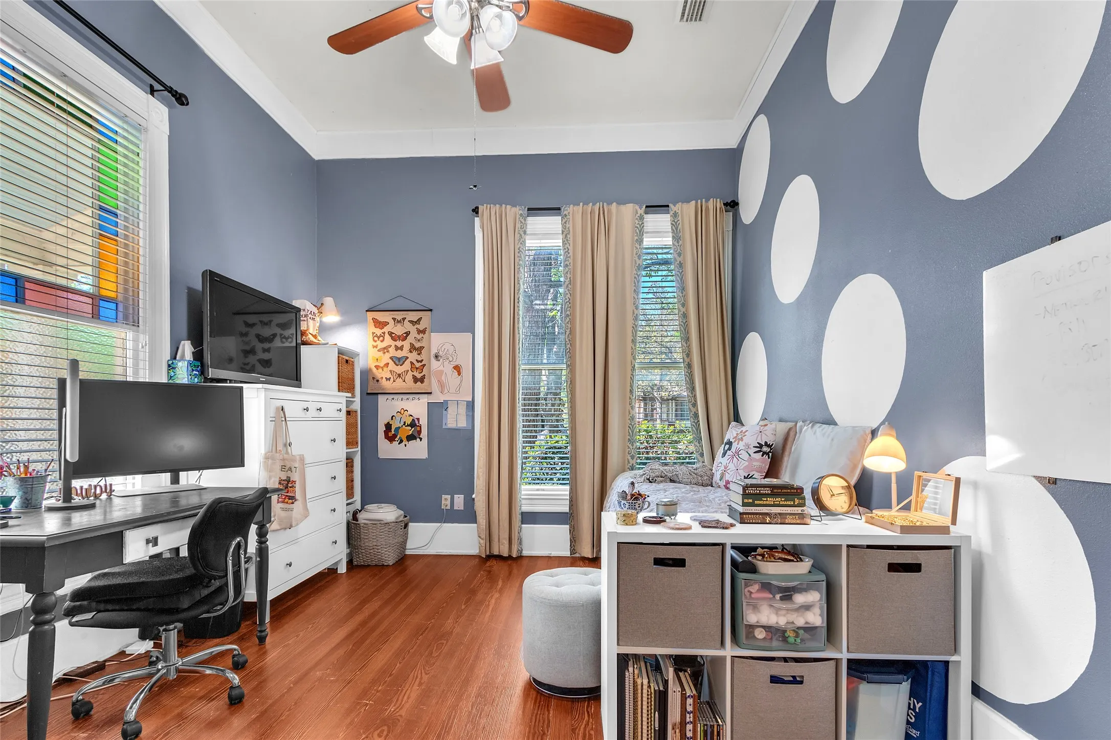 Cozy bedroom with hardwood floors, light blue walls adorned with white polka dots, large windows for natural light, and a functional layout including a work desk and storage unit