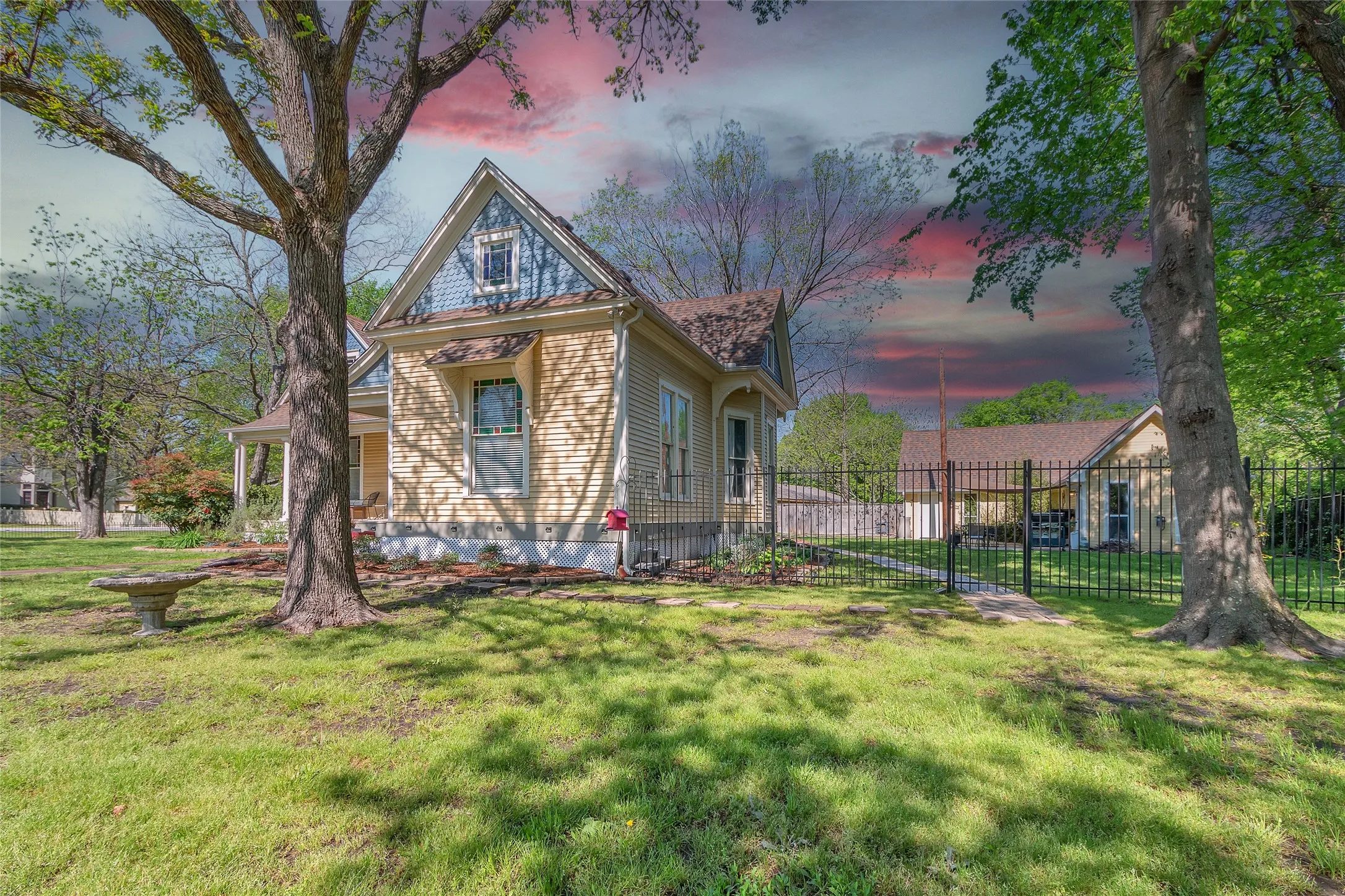 Well-maintained front yard with manicured grass, decorative shrubs, and a wooden bench adjacent to a two-story Victorian-style house under a colorful sky