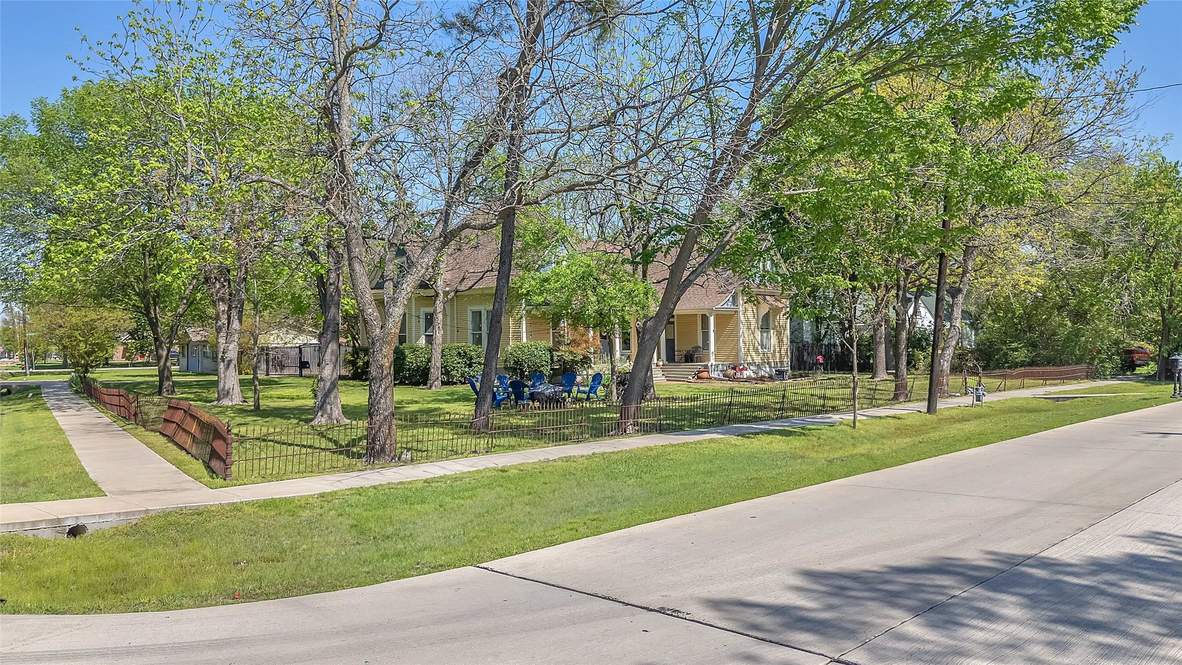 Front yard with manicured lawn, mature trees, decorative wrought iron fence, and path leading to a yellow house with a large covered porch