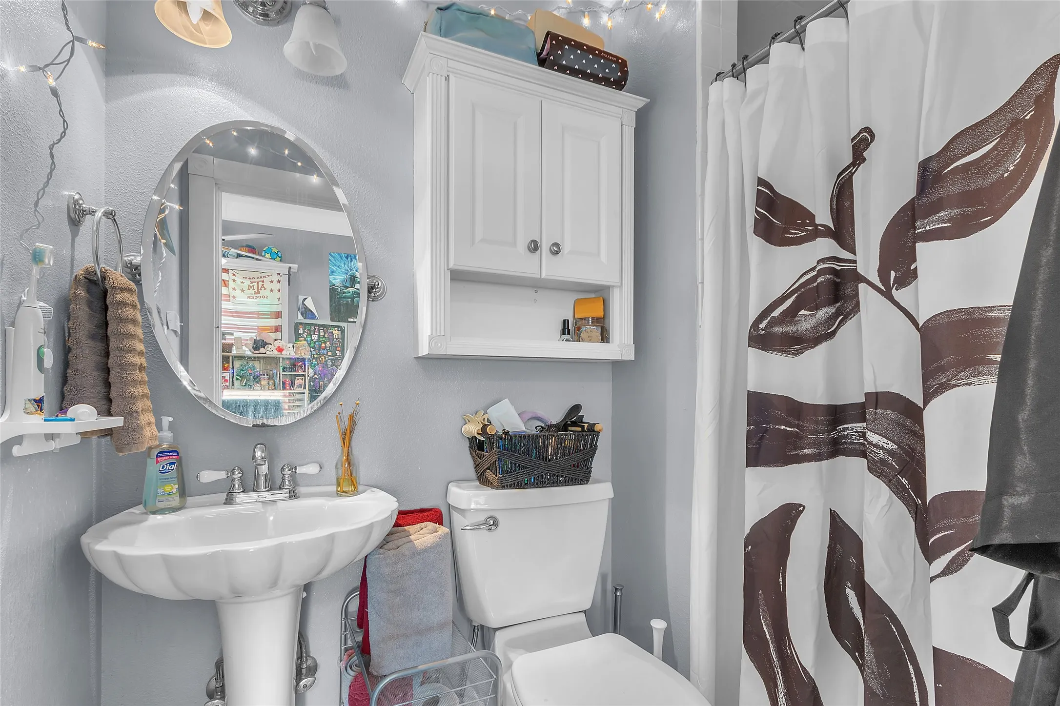 Bathroom with a pedestal sink, toilet, white cabinetry, shower curtain featuring leaf motif, and decorative string lights creating ambient lighting