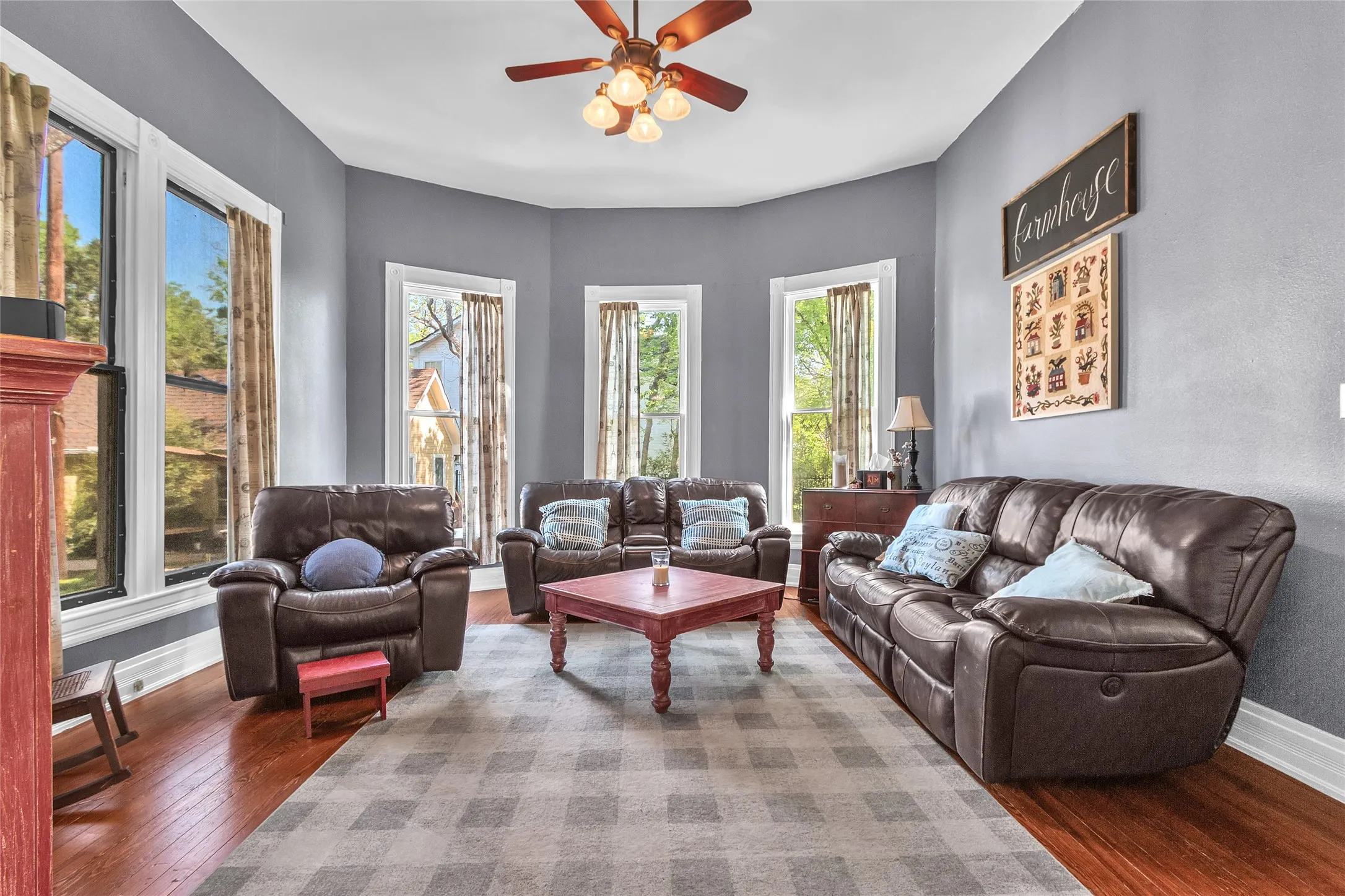 Living room with leather sofas, hardwood flooring, large bay windows allowing natural light, and a central coffee table in a cozy layout.