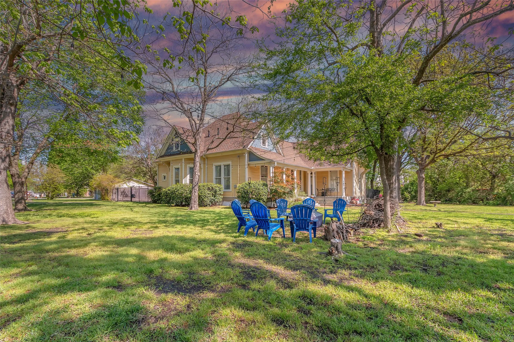 Residential exterior with an inviting yard, large grassy area, and blue Adirondack chairs set beneath mature trees near a two-story home surrounded by landscaping