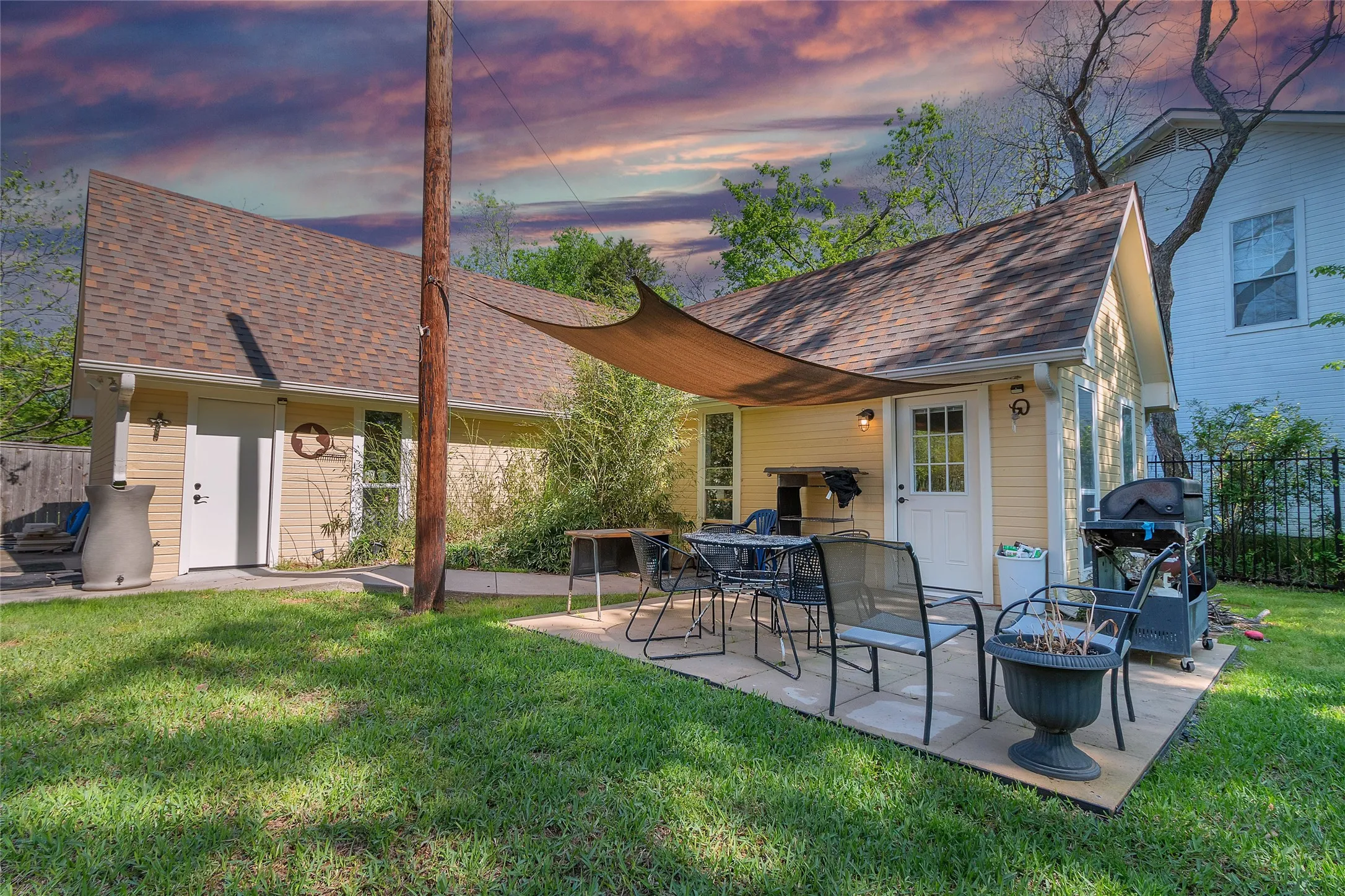 Covered back patio with outdoor dining set, stone pavers, and a grill under a shade sail amidst landscaped greenery and evening sky colors