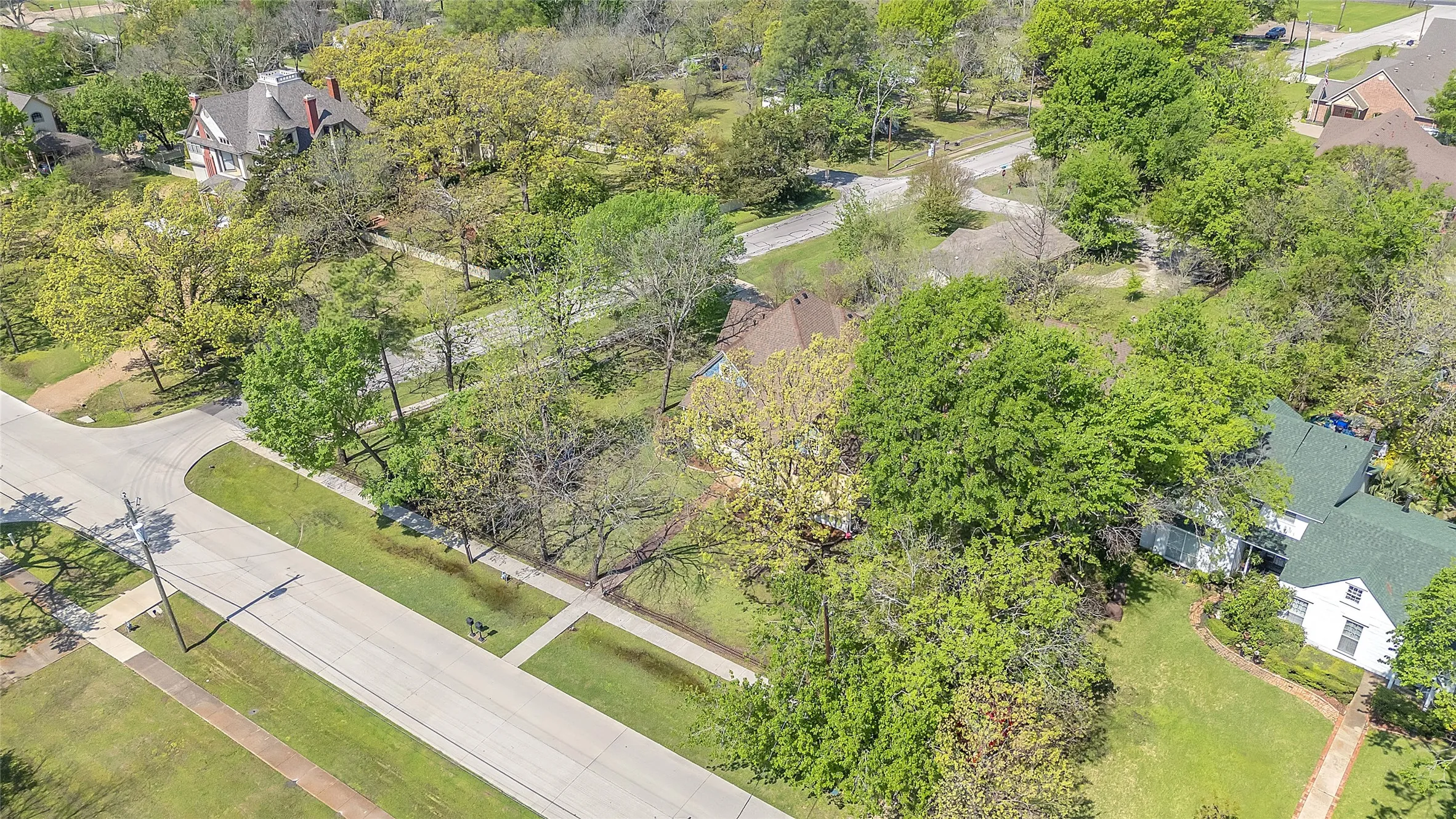 Aerial view of residential landscape showing mature trees, manicured lawns, and nearby streets with a mix of greenery and homes in a suburban setting