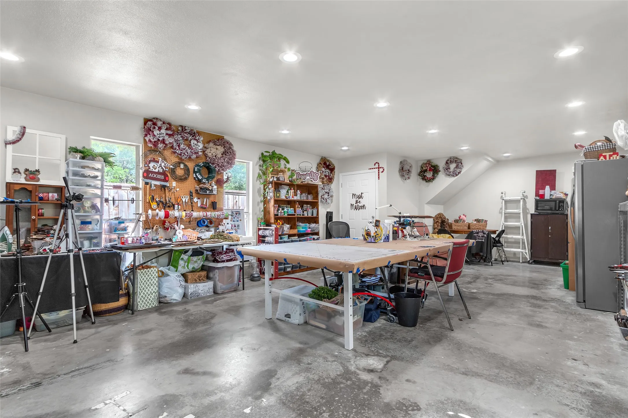 Workshop with various workstations, shelving units, and a large central table on a concrete floor, well-lit by recessed lighting with an open layout.