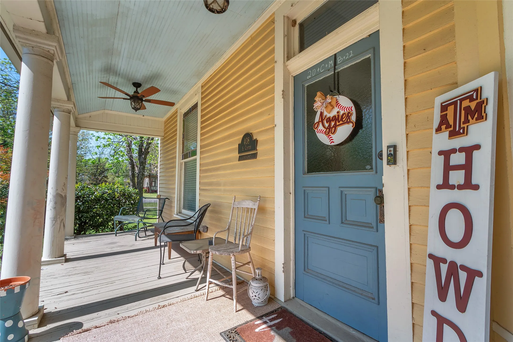 Covered front porch with wood flooring, ceiling fan, and decorative door wreath, complemented by a rustic rocking chair and potted​​‌​​​​‌​​‌‌​‌‌​​​‌‌​‌​‌​‌​​​‌​​ plants.