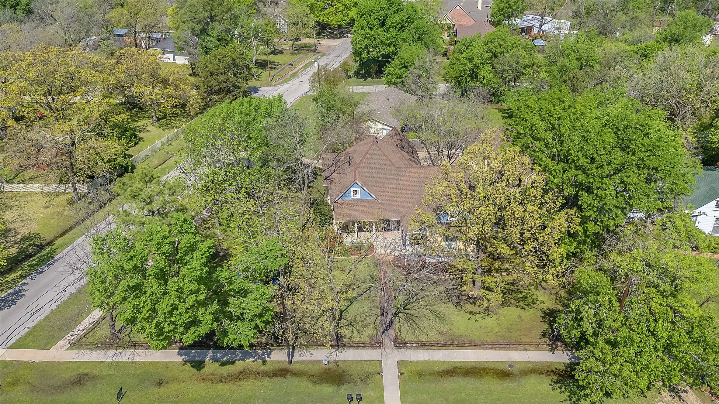 Aerial view of residential property with lush greenery, mature trees, and a well-maintained lawn near a quiet street and neighborhood homes.