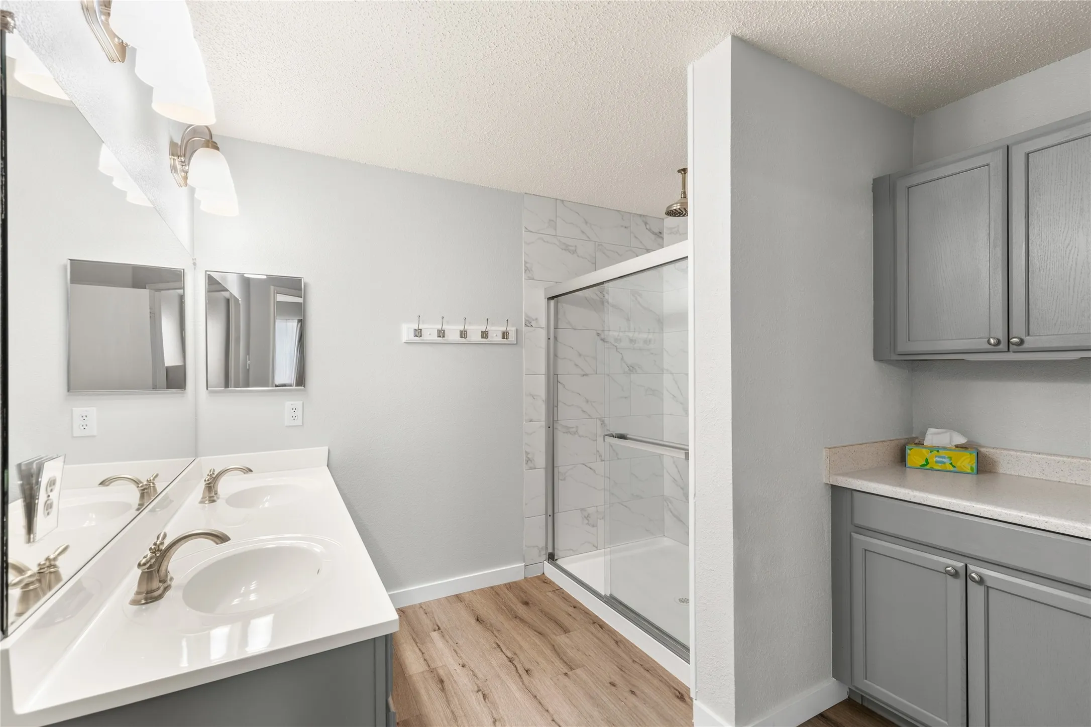Dual vanity with undermount sinks, wood-finish flooring, glass-enclosed shower with tile surround, and built-in cabinetry with counter space