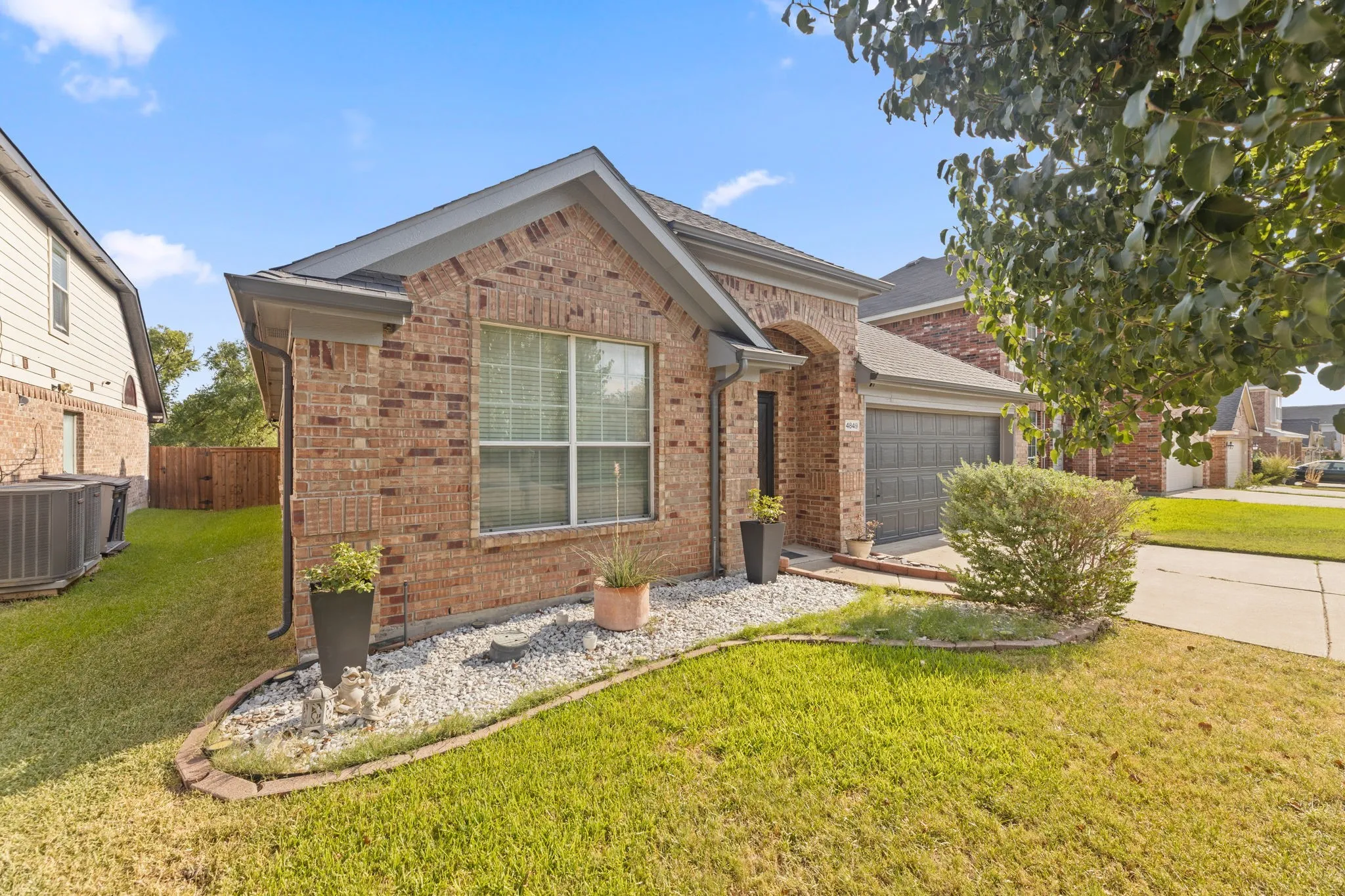 Charming brick single-story home featuring a manicured front lawn, mature landscaping with established shrubs, decorative gravel beds, and a two-car garage
