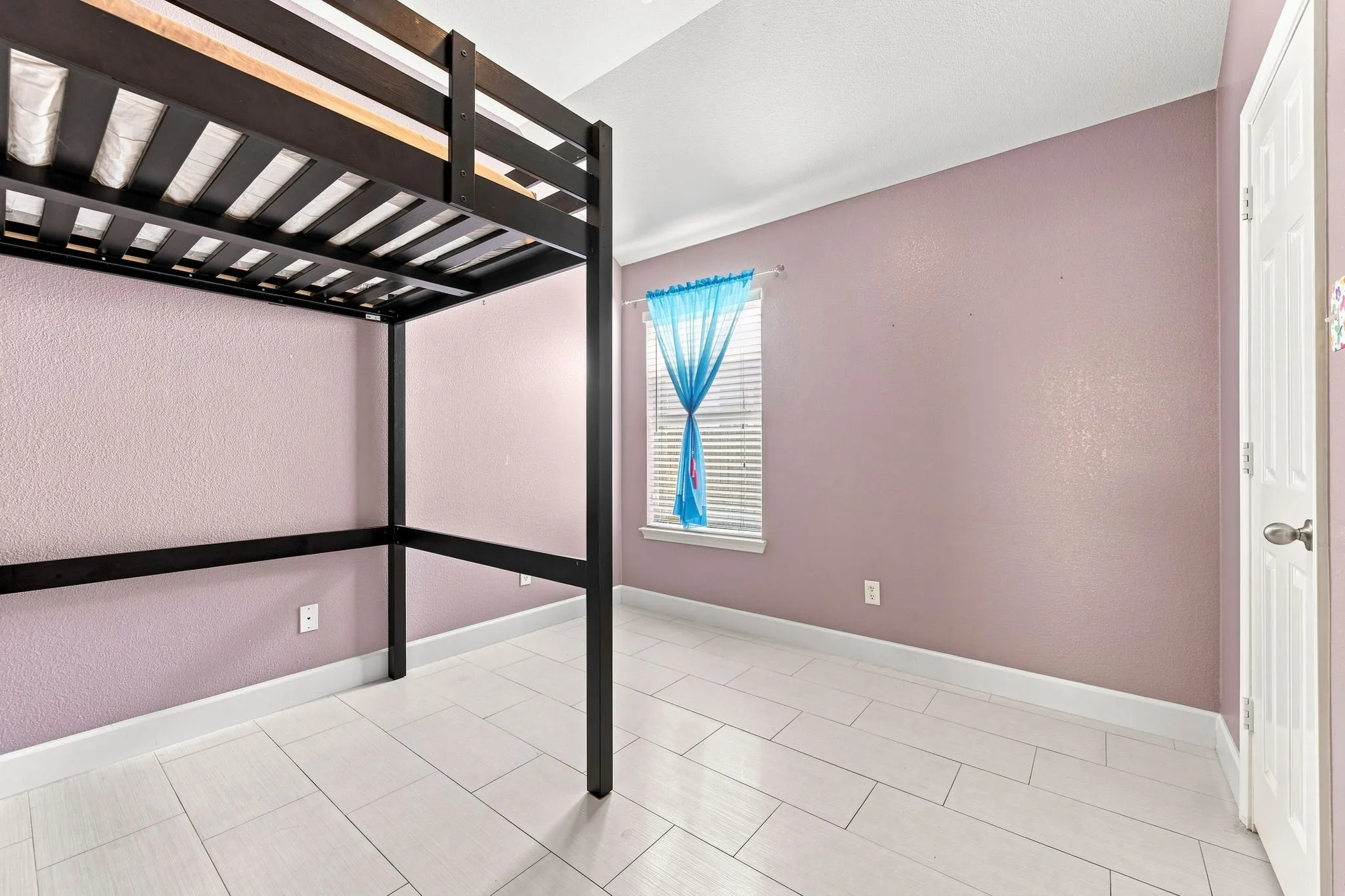 Spacious bedroom featuring a black metal loft bed frame with slatted support, ceramic tile flooring, mauve walls, and abundant natural light from a window with blue drapery