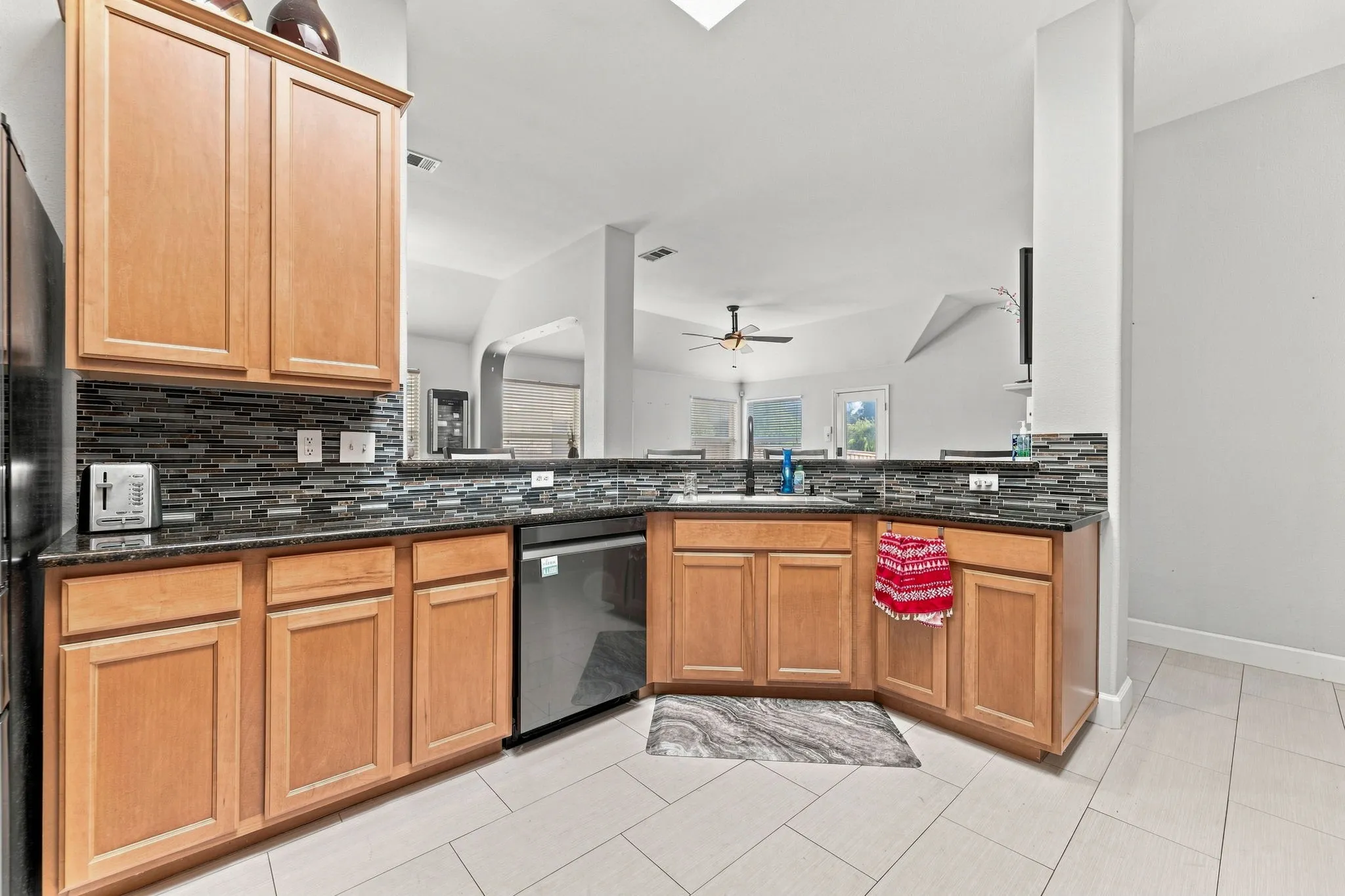 Modern kitchen featuring warm wood cabinetry, dark tile backsplash, stainless steel appliances, granite countertops, and ceramic tile flooring