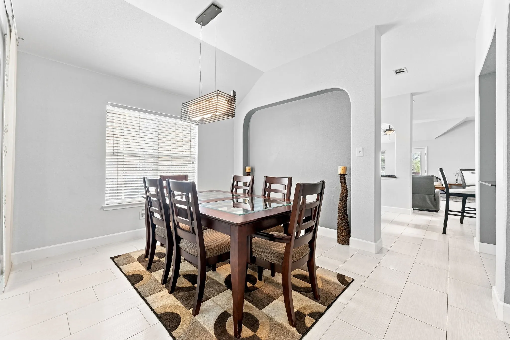 Vaulted ceiling with a contemporary pendant light fixture, window with horizontal blinds, arched wall opening, and light-toned tile flooring