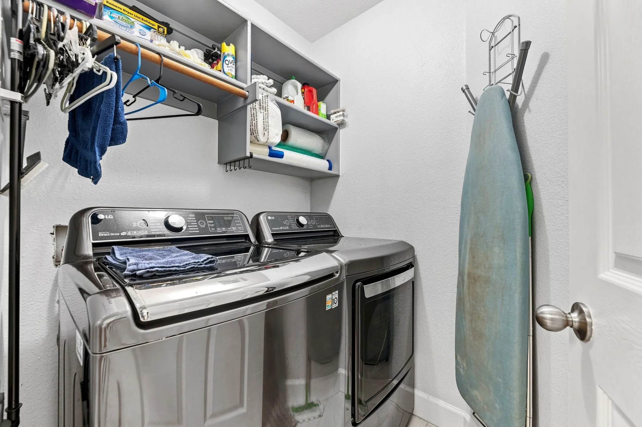Dedicated laundry room featuring side-by-side stainless steel washer and dryer with modern digital controls, overhead shelving for storage and supplies, wall-mounted drying rack, and direct door access