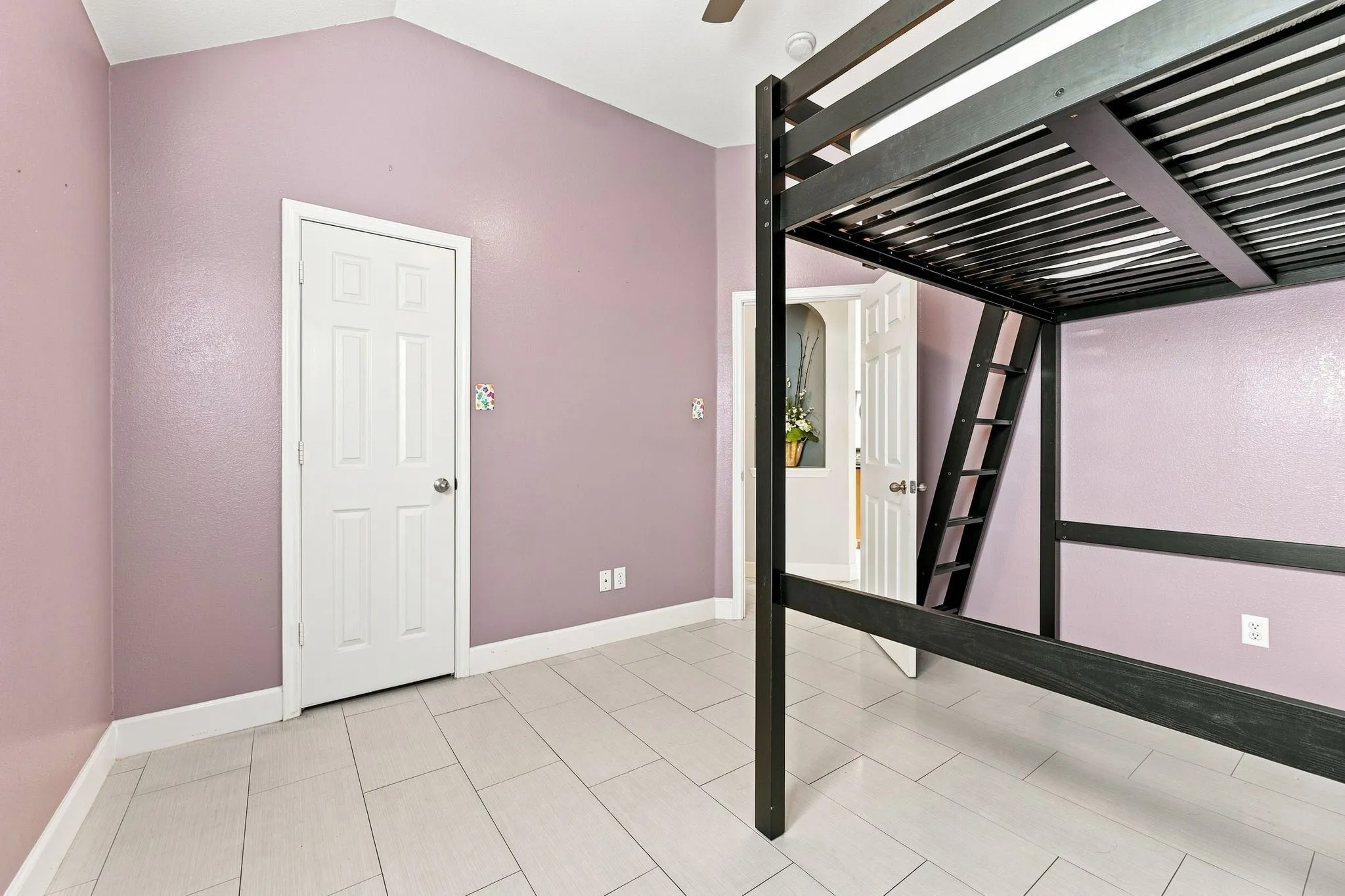 Spacious bedroom with soft mauve walls, ceramic tile flooring, white six-panel entry door, and a black metal loft-style bed frame with integrated ladder and overhead storage structure