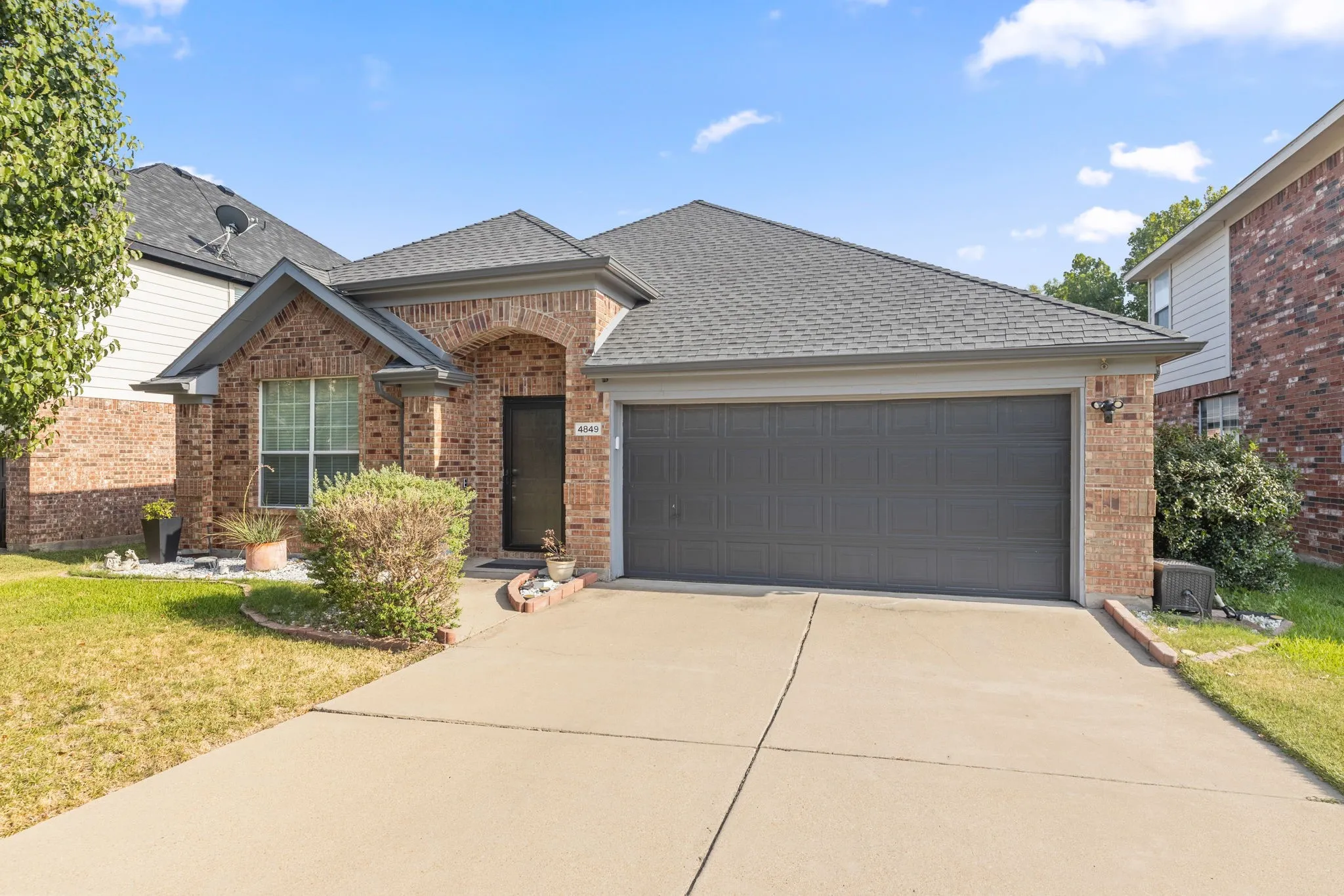 Charming brick home featuring a spacious driveway, dark gray garage door with modern paneling, arched entryway detail, and lush landscaping with mature foundation plantings