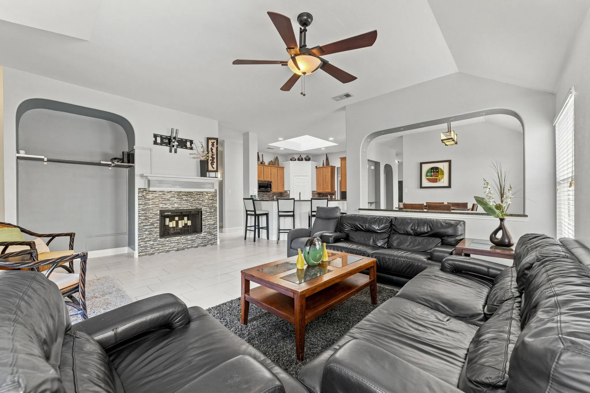 Open-concept living area featuring a fireplace with stone surround and mantel display, ceiling fan with integrated lighting, wood-finish coffee table, black leather sectional seating, and multiple architectural arches with rounded frames throughout the space