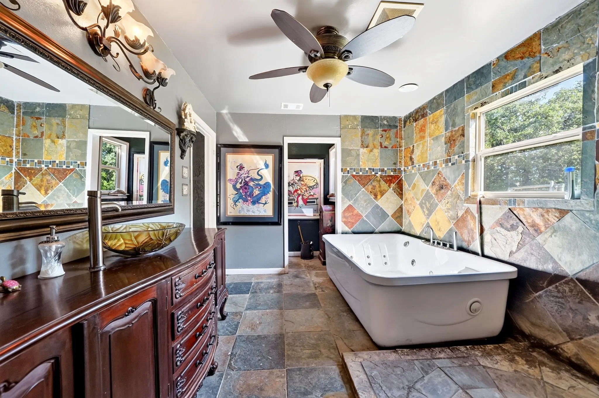 Spa-inspired primary bathroom showcasing a freestanding soaking tub, dual vanity with wood cabinetry and integrated sink, decorative tile accents throughout, ceiling fan, and abundant natural light from multiple windows