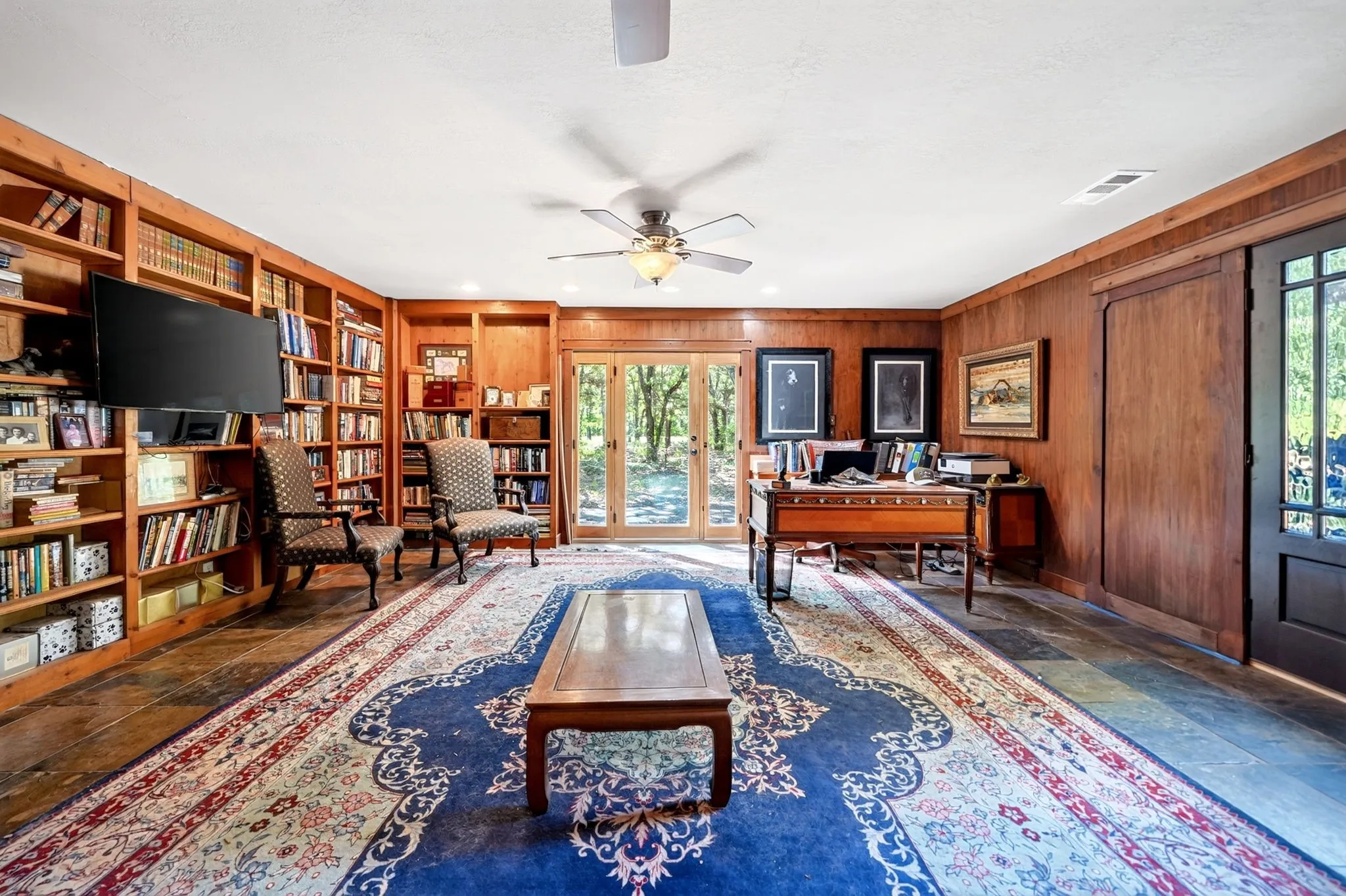 Elegant wood-paneled study featuring extensive built-in shelving, a ceiling fan with brass accents, hardwood flooring, and French doors with transom windows overlooking landscaped grounds