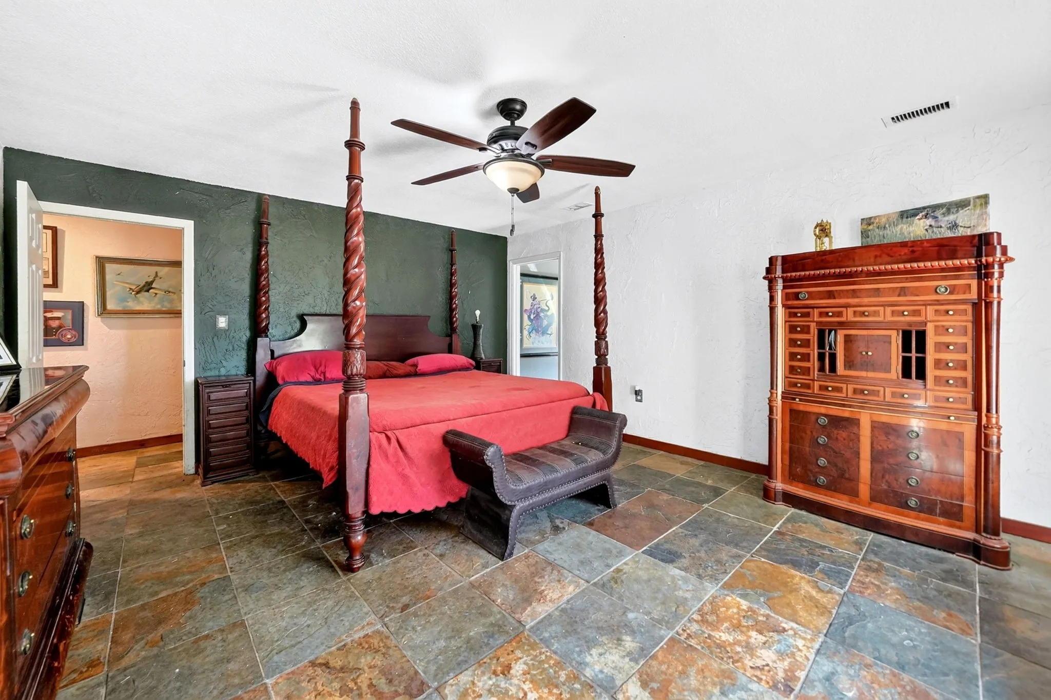 Primary bedroom suite featuring a four-poster bed frame, slate-finish tile flooring in a geometric pattern, sage green accent walls, ceiling fan with integrated lighting, and wood-finish cabinetry