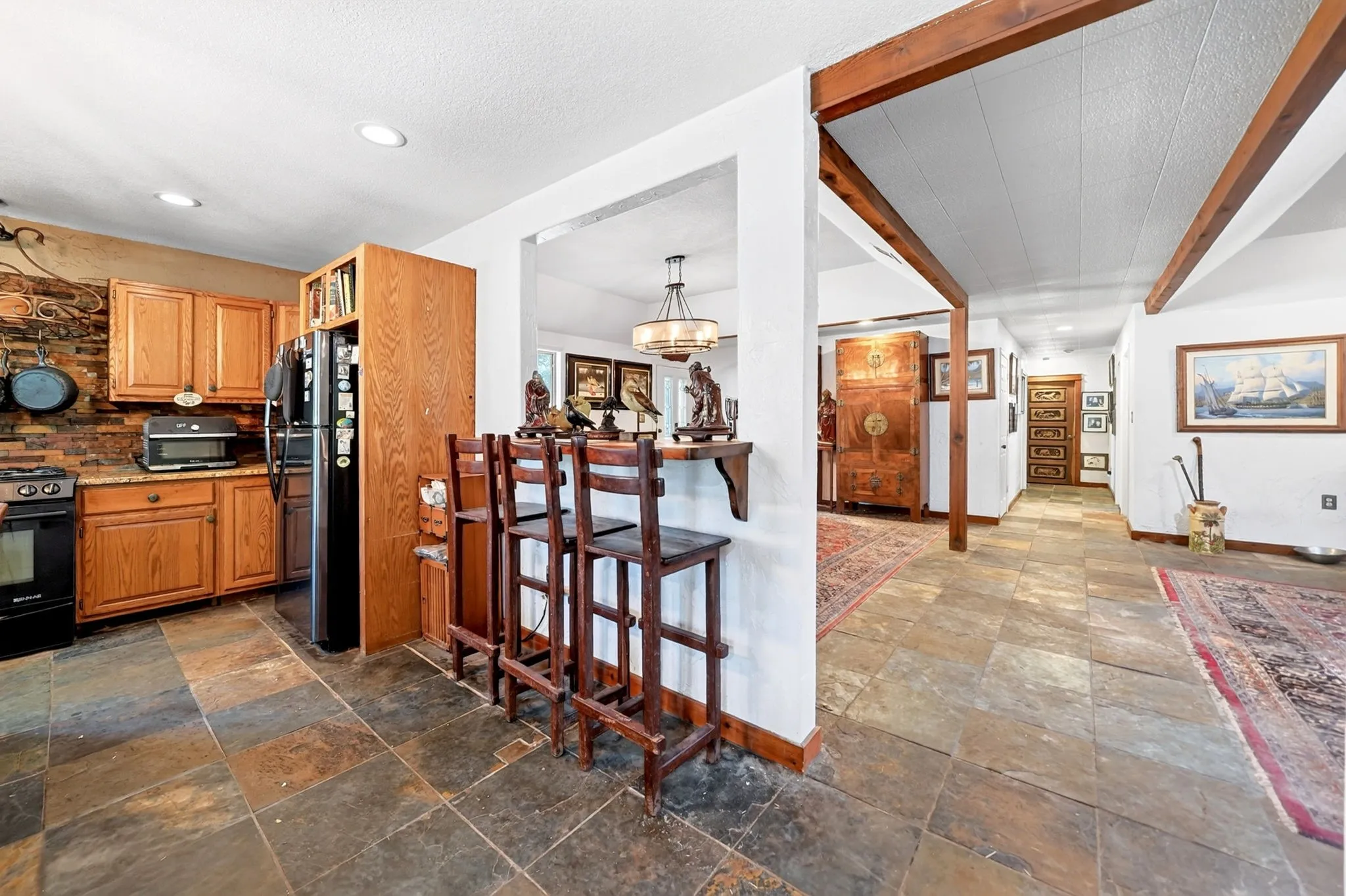 Open-concept kitchen and dining area featuring wood cabinetry, stainless steel appliances, slate flooring, exposed beam ceiling details, and a central island with seating