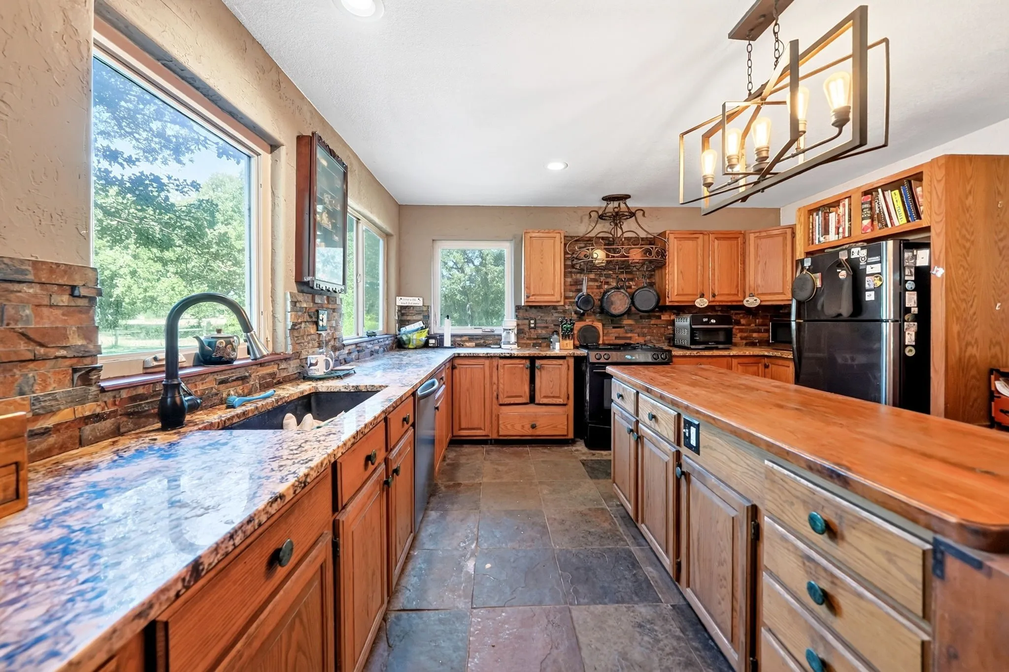 Chef's kitchen featuring warm wood cabinetry, multi-toned stone countertops, black stainless steel appliances, a large center island with wood-finish countertop, and abundant natural light from expansive windows