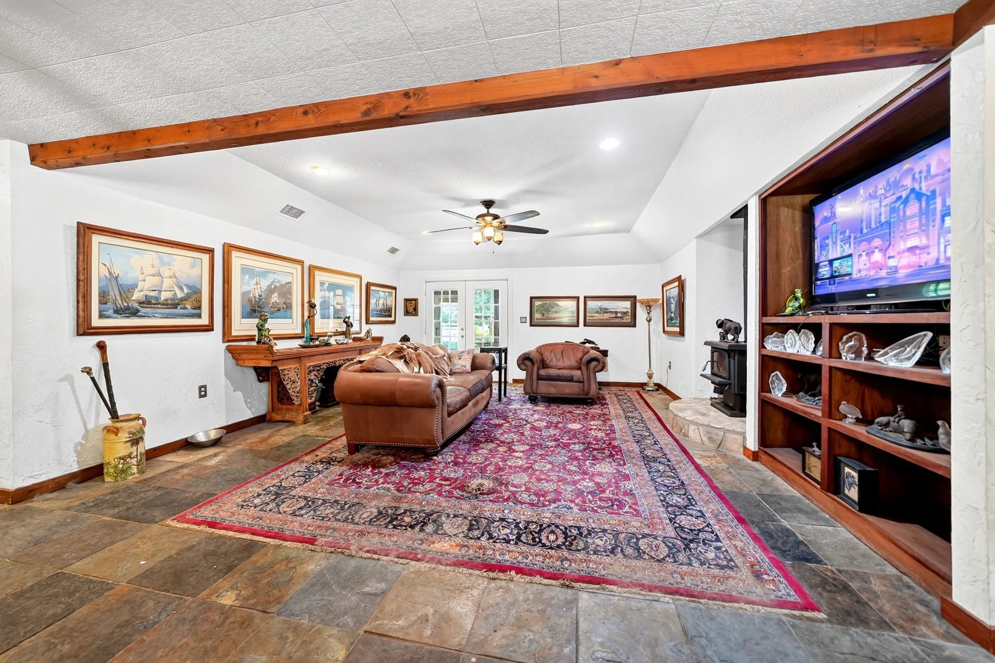 Spacious recreation room showcasing exposed timber beam ceiling detail, stone-finish flooring, built-in wood cabinetry with shelving, ceiling fan, and recessed lighting