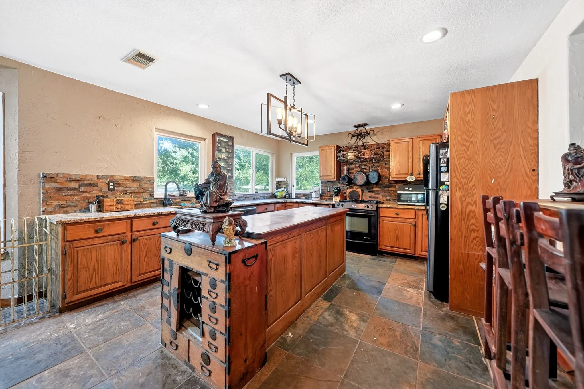 Spacious kitchen featuring warm wood cabinetry, a large center island with seating, stainless steel appliances, tile flooring, abundant natural light from multiple windows, and recessed ceiling lighting