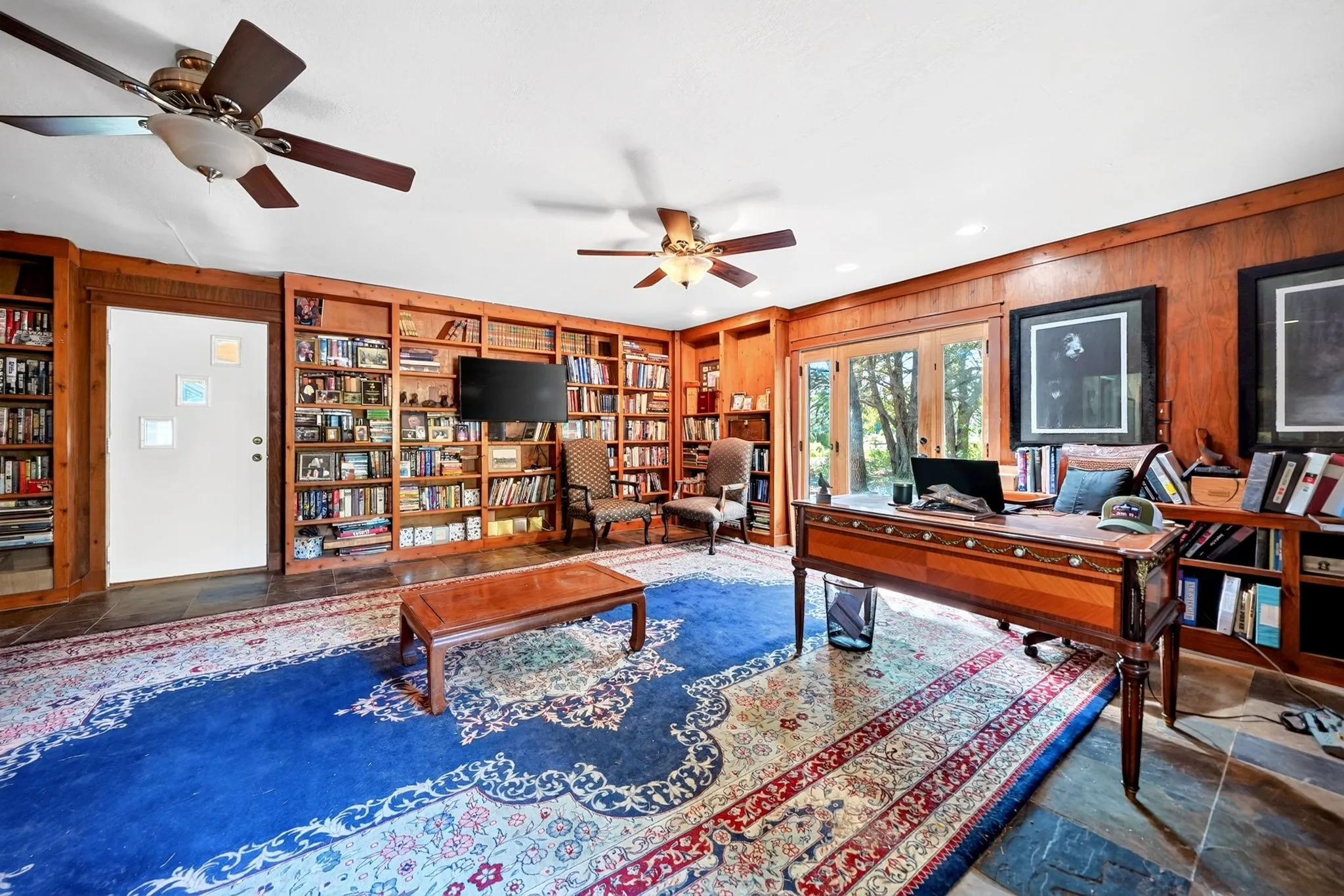 Wood-paneled library featuring extensive built-in shelving, two ceiling fans, multiple windows with natural light, and hardwood-finish flooring anchored by layered area rugs
