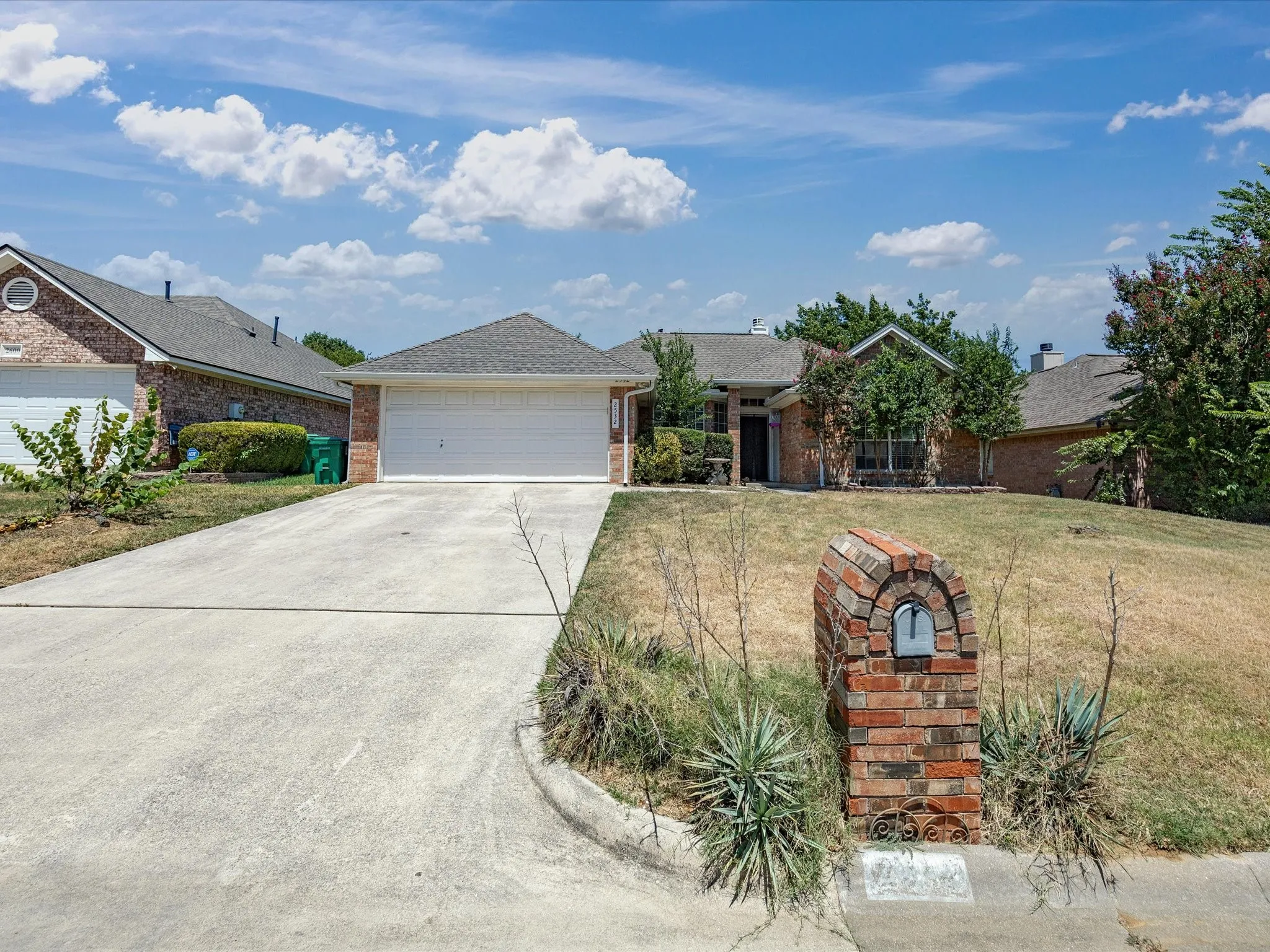 Brick exterior with an attached garage, concrete driveway, and mature landscaping