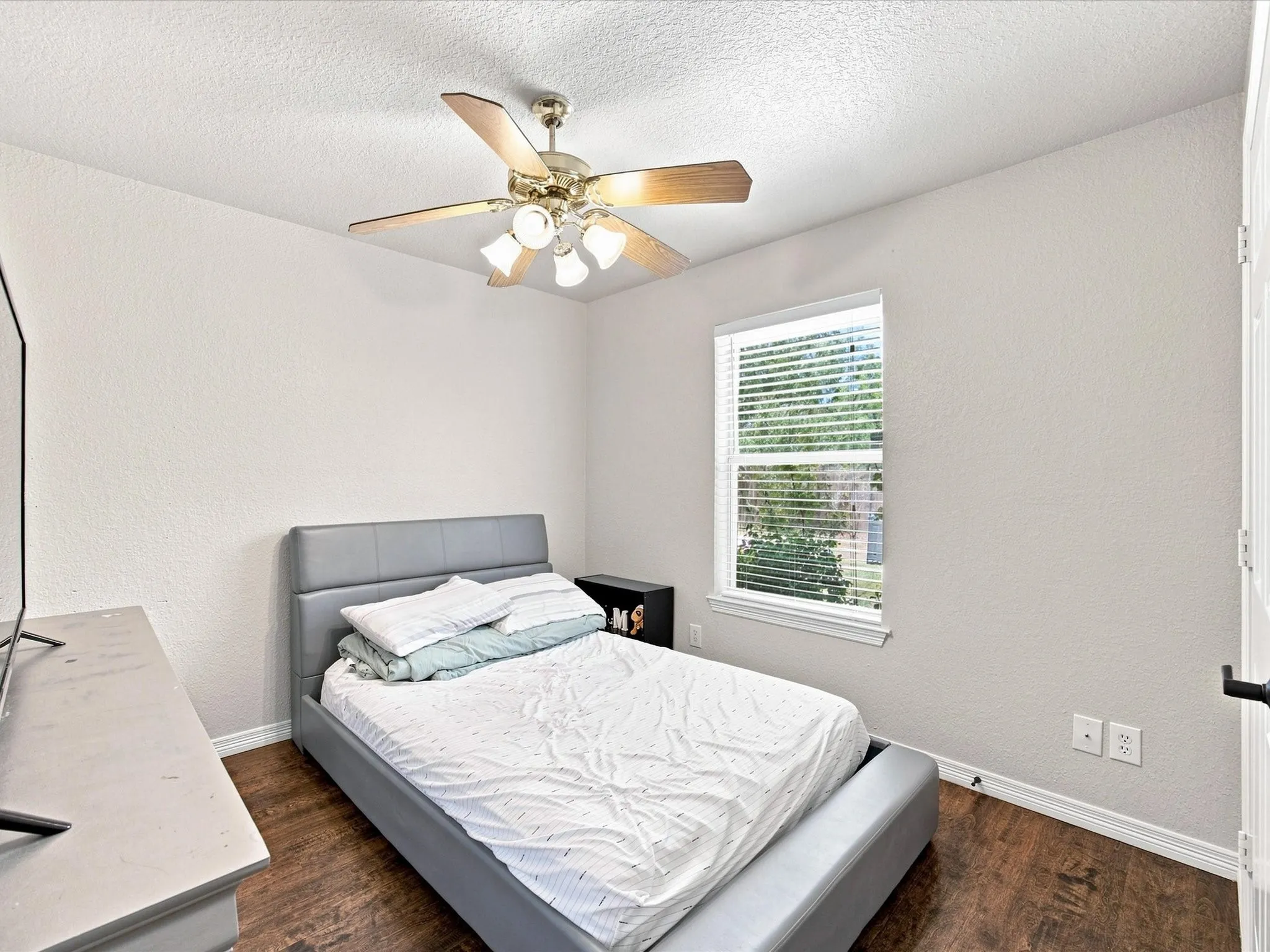 Wood-finish flooring, single window with blinds, ceiling fan with integrated lighting, textured walls, and a white interior door
