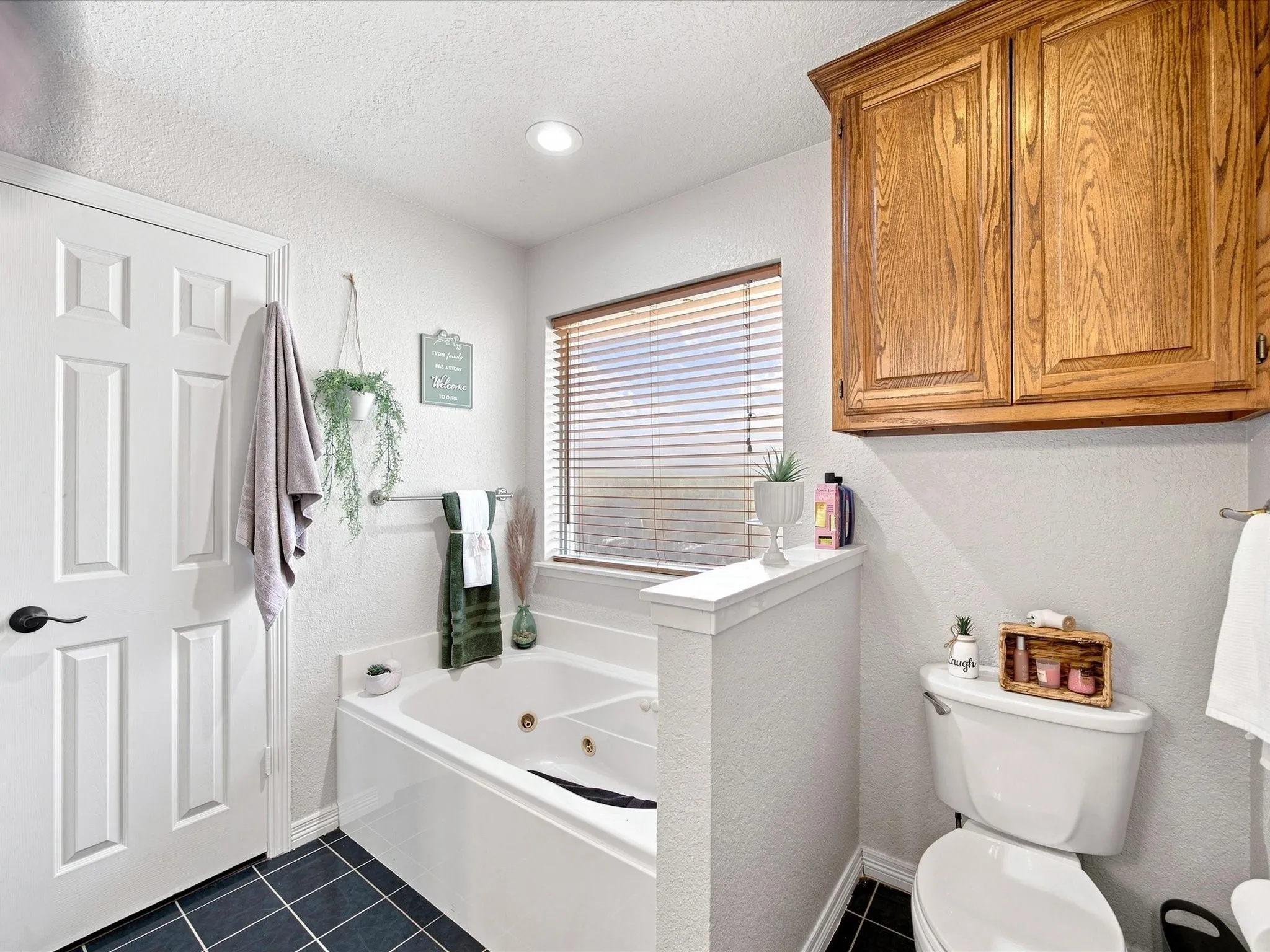 Jetted soaking tub with a privacy wall, window with blinds, overhead wood cabinetry, black tile flooring, and a white toilet