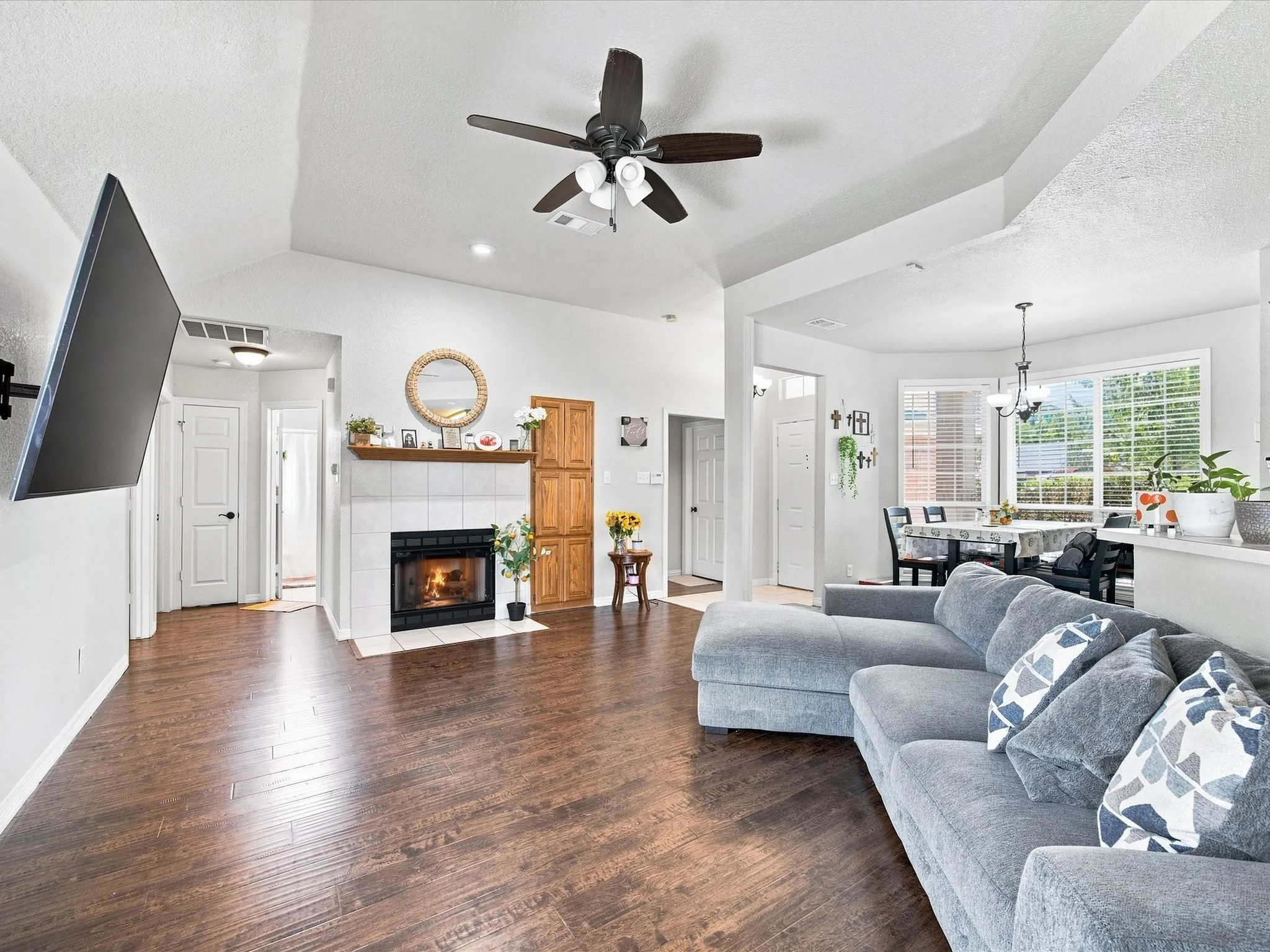 Vaulted ceilings with a ceiling fan, a tiled fireplace with a wood mantle, wood-finish flooring, and white wall​​‌​​​​‌​​‌‌​‌‌​​​‌‌​‌​‌​‌​​​‌​​ finishes