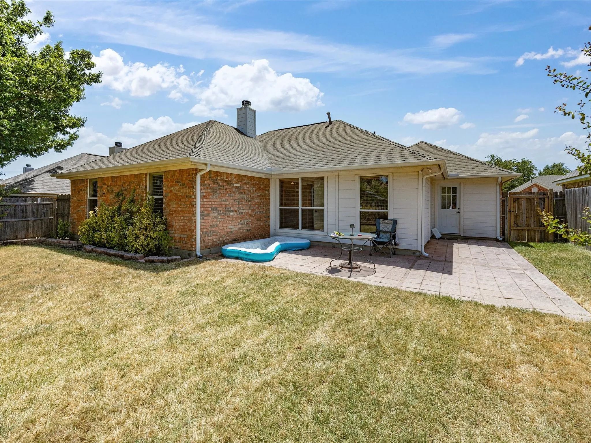 Exterior brick and siding combination, paved patio area, fenced perimeter, multiple windows, and a mature tree