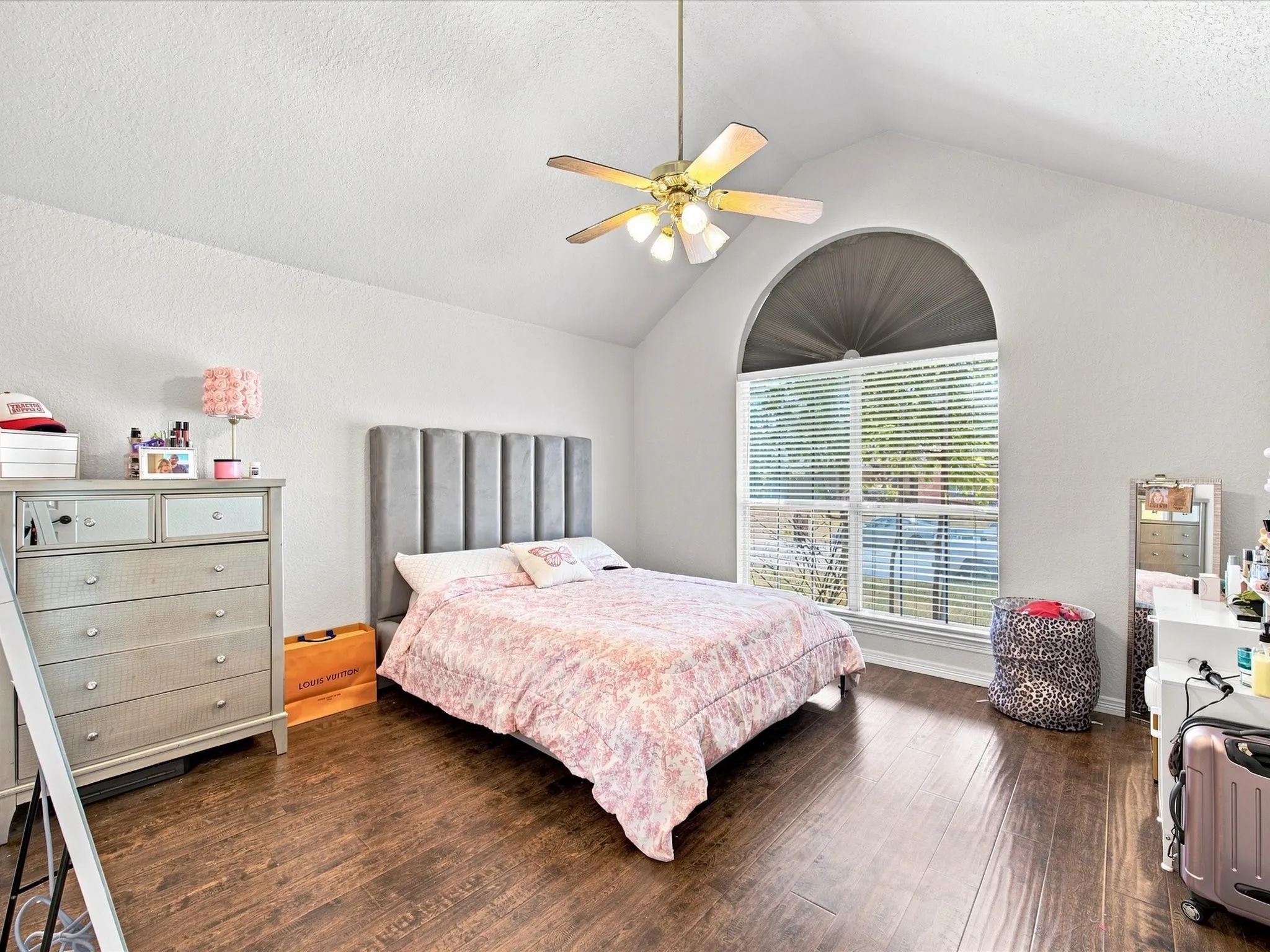 Spacious primary bedroom featuring vaulted ceilings with integrated ceiling fan and recessed lighting, arched transom window with plantation shutters, wood-finish flooring, and coordinating dresser with built-in storage