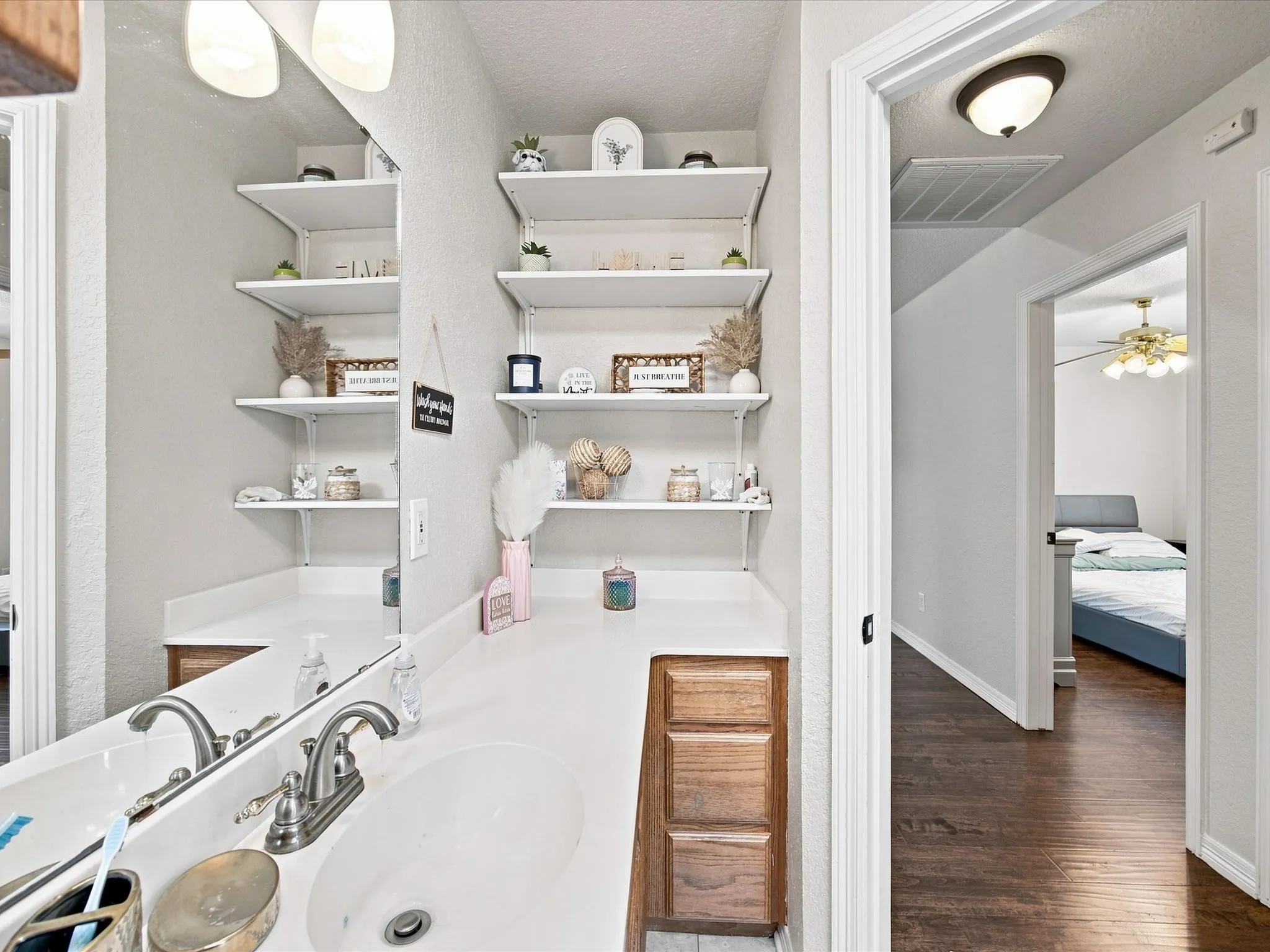 Bright primary bathroom featuring dual vanity with brushed nickel faucetry and white countertop, abundant floating shelving with decorative storage, recessed and ceiling-mounted lighting, wood-finish flooring, and direct access to adjoining bedroom space