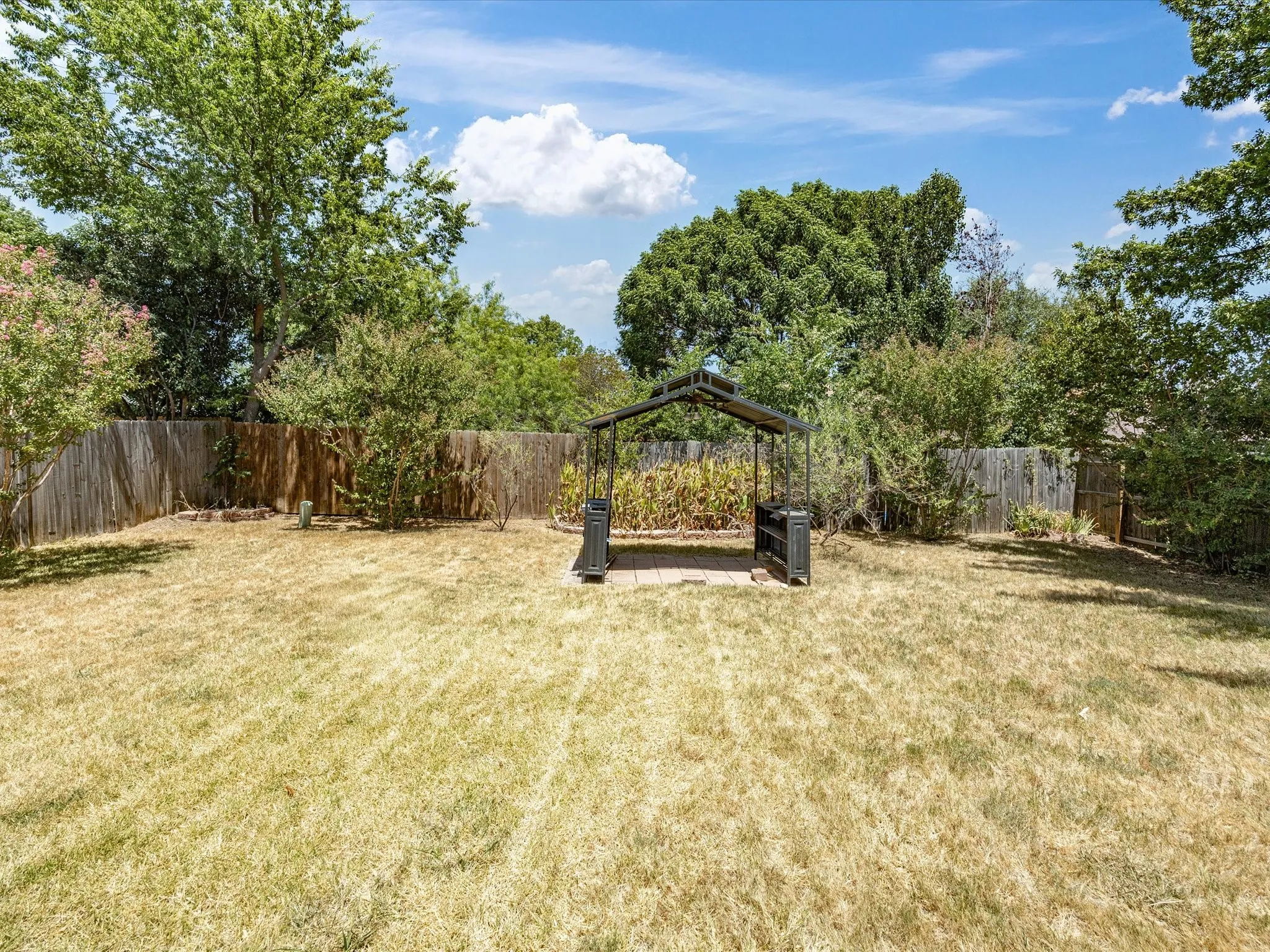 Expansive backyard featuring a privacy fence, mature trees, and a covered patio area with a paved surface