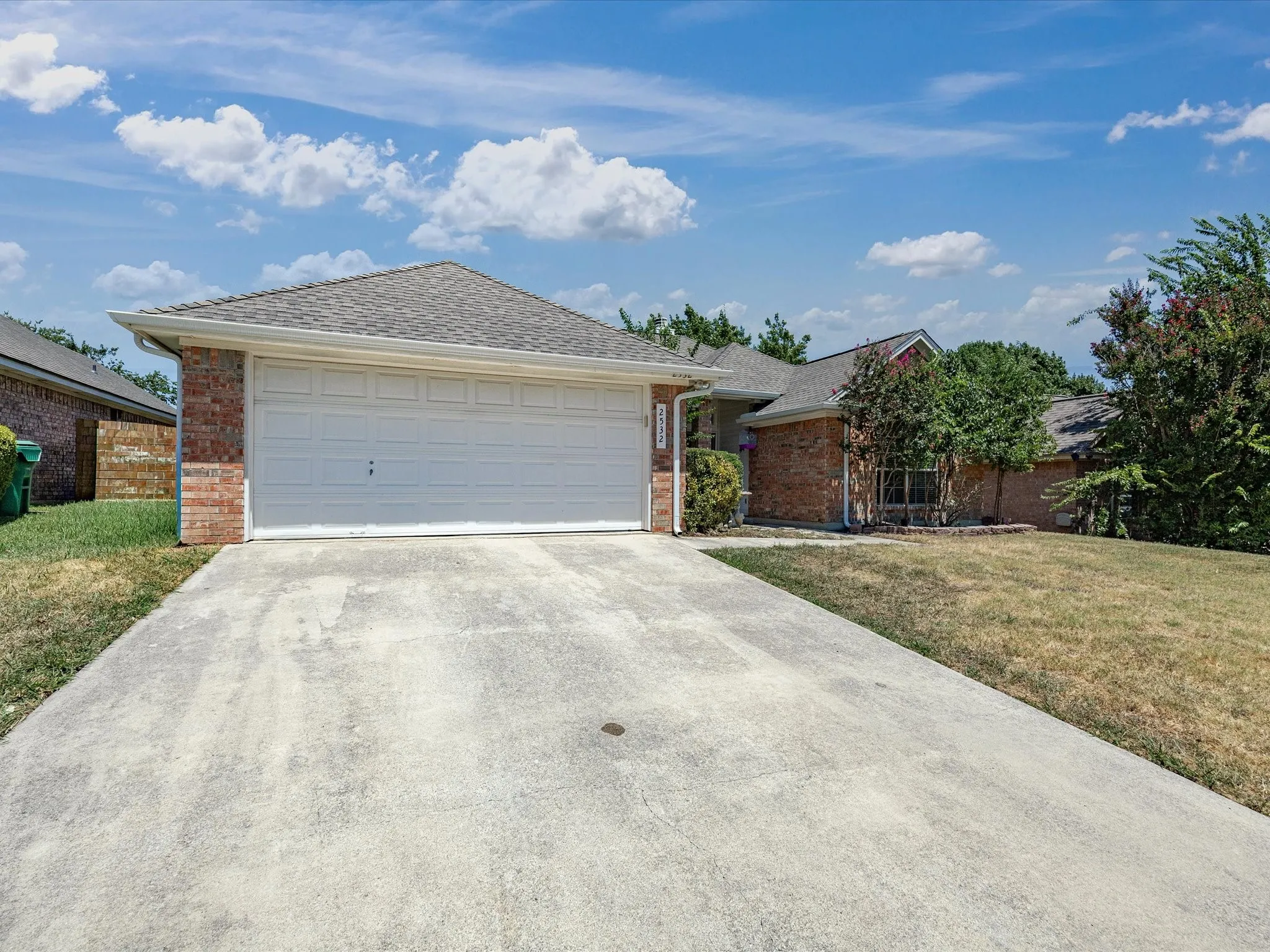 Charming brick home featuring a spacious two-car garage with white paneled doors, concrete driveway, and mature landscaping