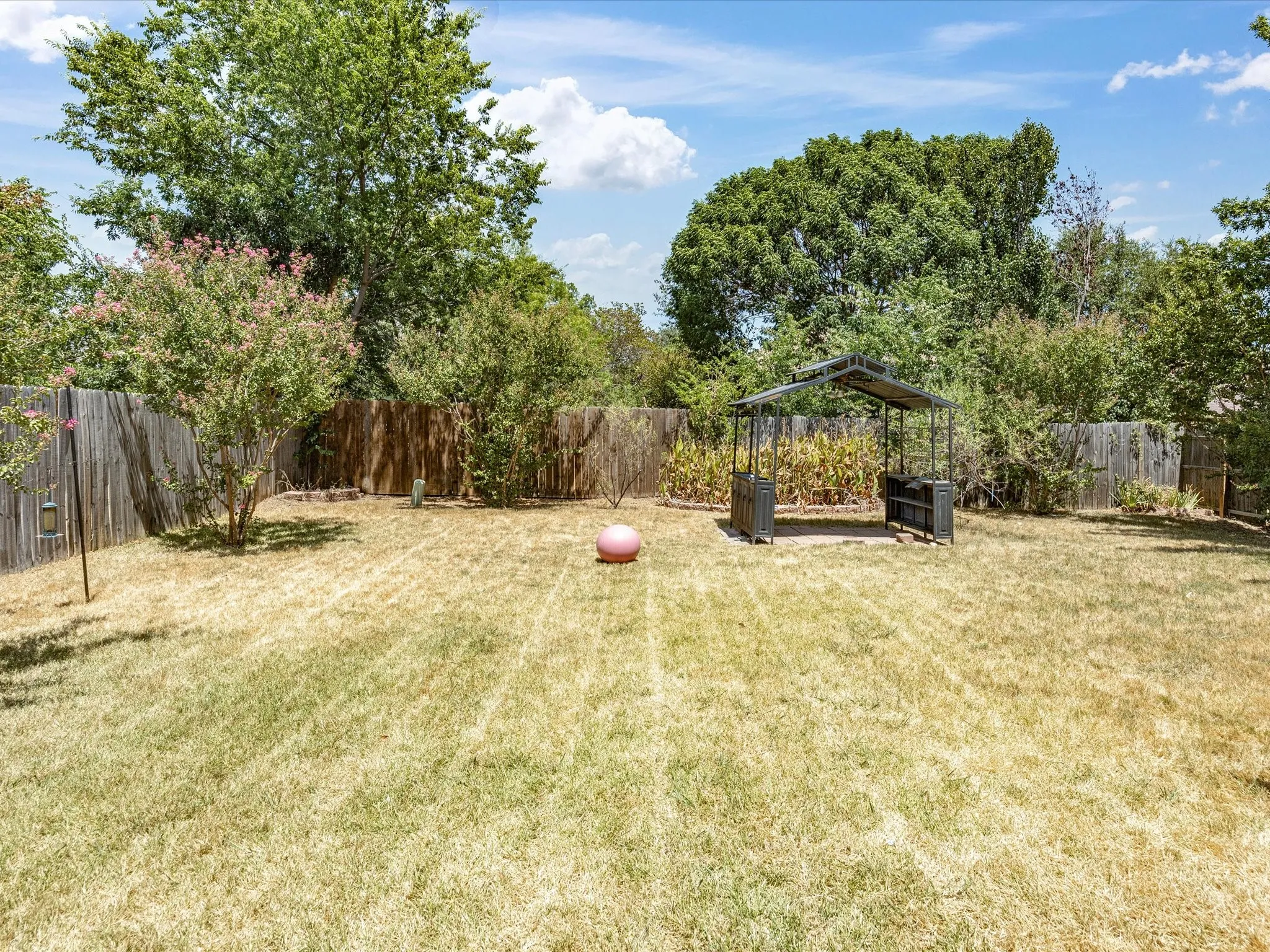 Spacious backyard oasis featuring mature shade trees, manicured lawn with defined striping, wooden privacy fencing, and multiple hardscape structures including a pergola and storage shed