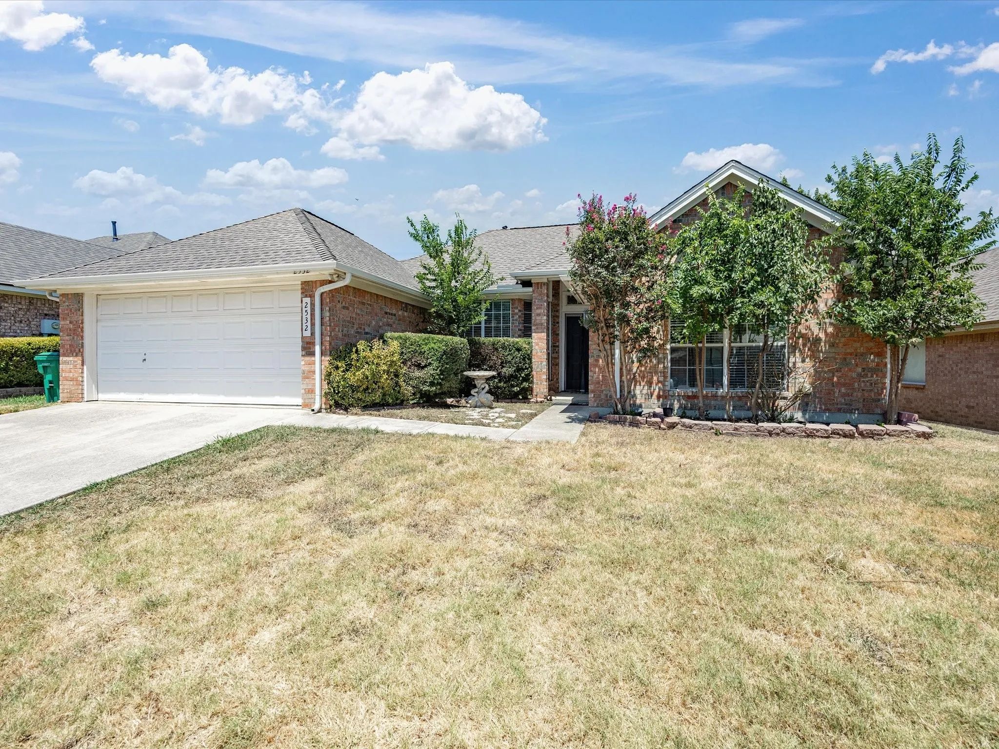 Charming single-story brick residence featuring a two-car garage with white paneled door, manicured front lawn, and mature landscaping including flowering shrubs and established trees