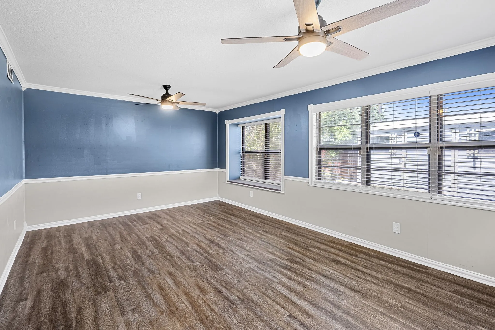 Wood-finish flooring throughout, multiple windows with blinds, two ceiling fans with integrated lighting, and crown molding