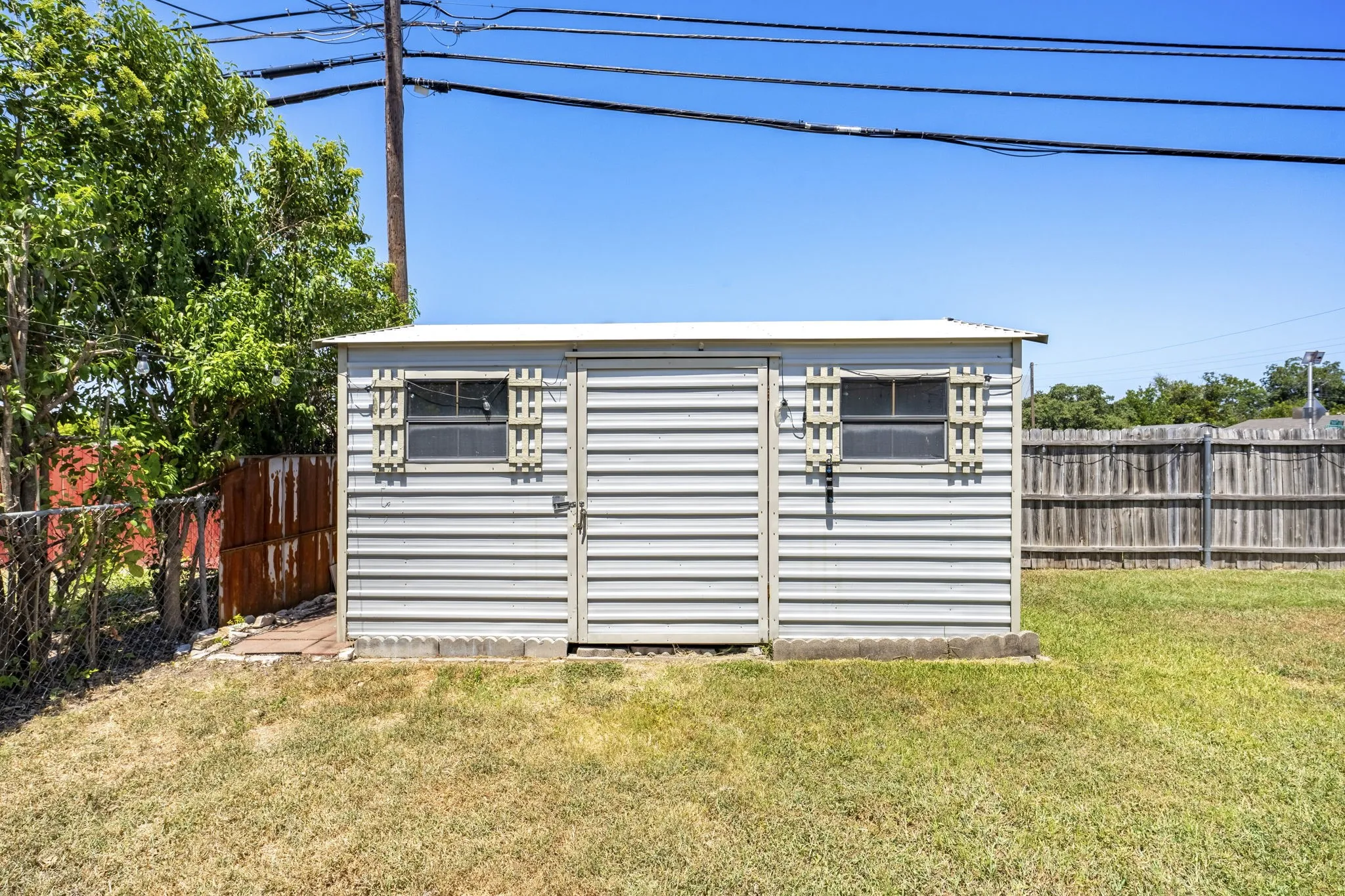 Detached storage shed with corrugated metal siding, two windows with decorative shutters, and a flat roof