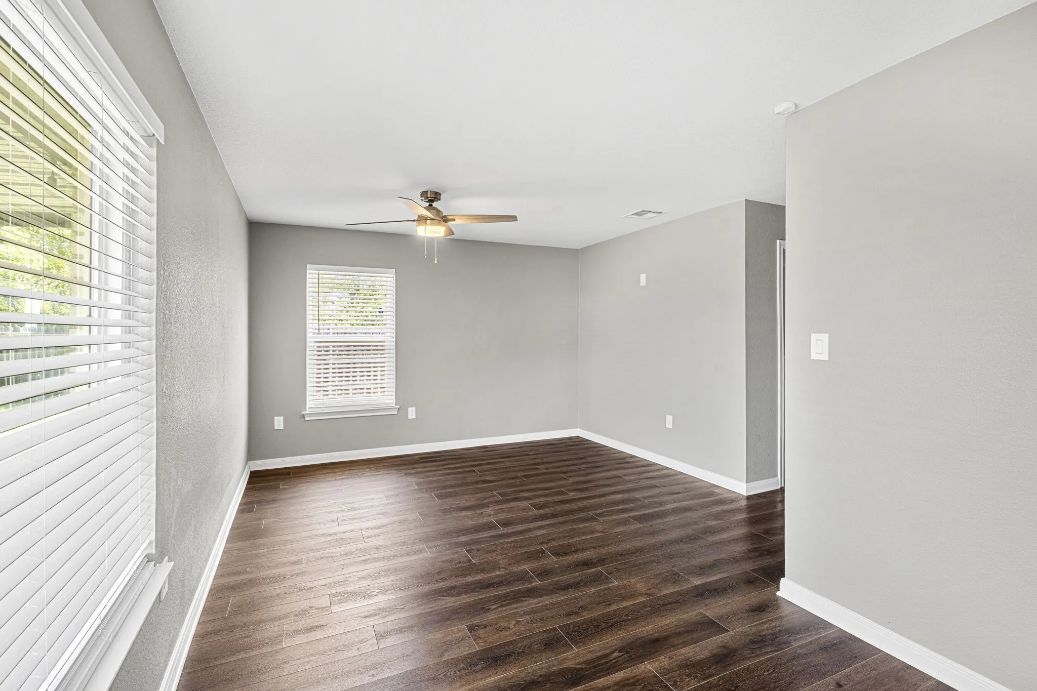 Spacious room with wood-finish flooring, two windows with blinds, a ceiling fan, and white trim
