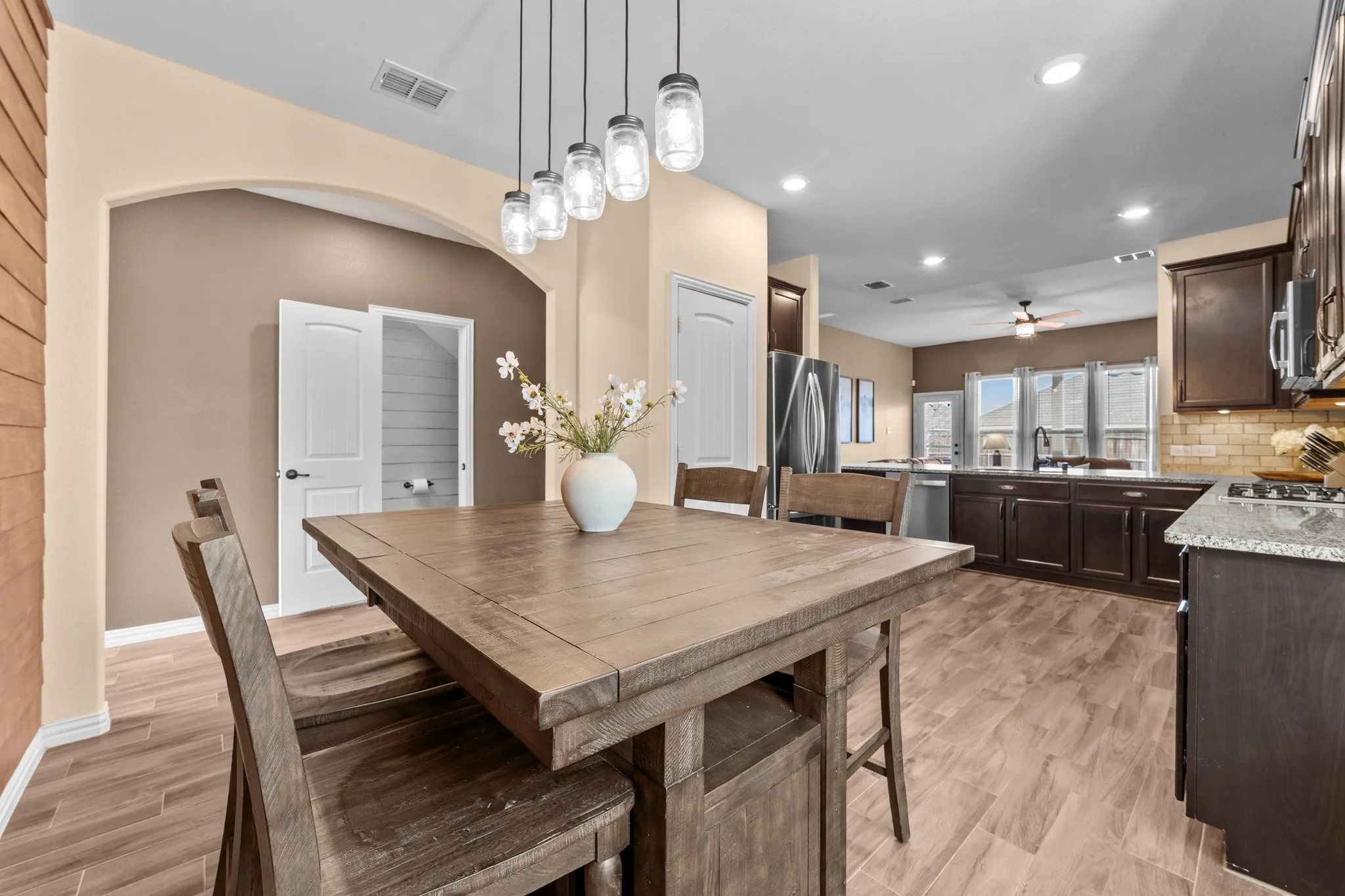 Open-concept dining and kitchen area showcasing wood-finish tile flooring throughout, pendant lighting with glass shades, stainless steel appliances, dark cabinetry with granite countertops, and recessed ceiling lighting