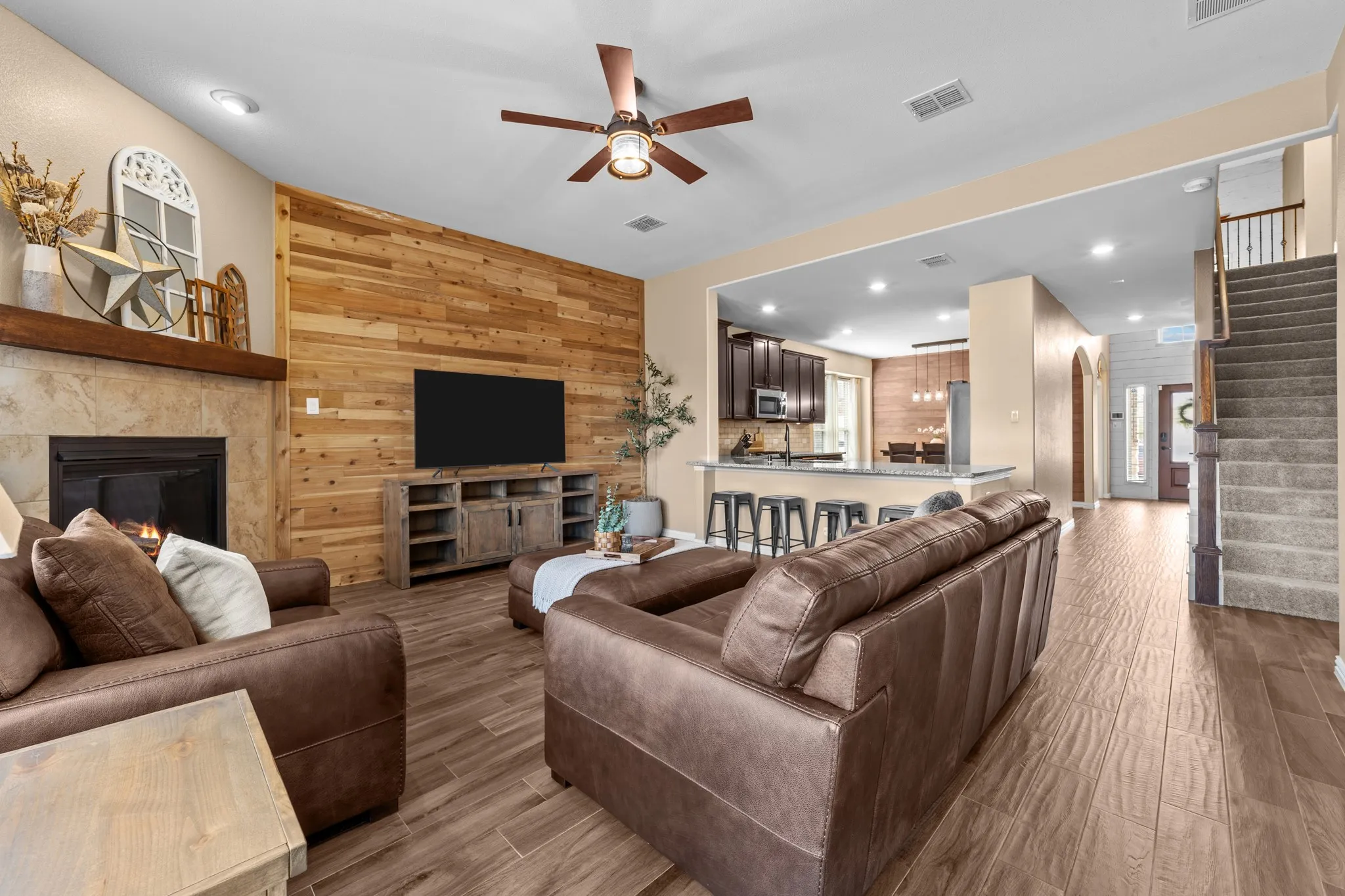 Open-concept living space featuring a wood-paneled accent wall, ceiling fan with integrated lighting, wood-finish tile flooring throughout, and a gas fireplace with surround